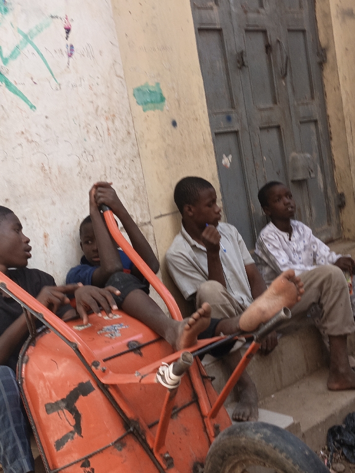 This image, taken in Kawaji, Nigeria, displays four young males in an outdoor urban setting. In the foreground, an orange metal wheelbarrow lies on its side, its single rubber wheel visible at the bottom right. A boy, wearing a blue shirt and dark trousers, holds the wheelbarrow's handles, with his bare feet resting on its inner frame. To his left, another boy in a dark shirt is partially visible, facing inward. To the right of the wheelbarrow, a third boy, dressed in a light-colored short-sleeved collared shirt, is seated against a beige wall marked with extensive green and other colored graffiti and stains; he appears to be holding a small dark object near his mouth. His bare feet are visible, resting on the ground near the wheelbarrow's framework. Further right, a fourth boy, wearing a white patterned long-sleeved shirt, is seated on concrete steps. The background features the aforementioned marked wall and a weathered, grey-blue double wooden door with visible metal hardware. A dark, possibly plastic, object is on the ground near the steps on the far right.