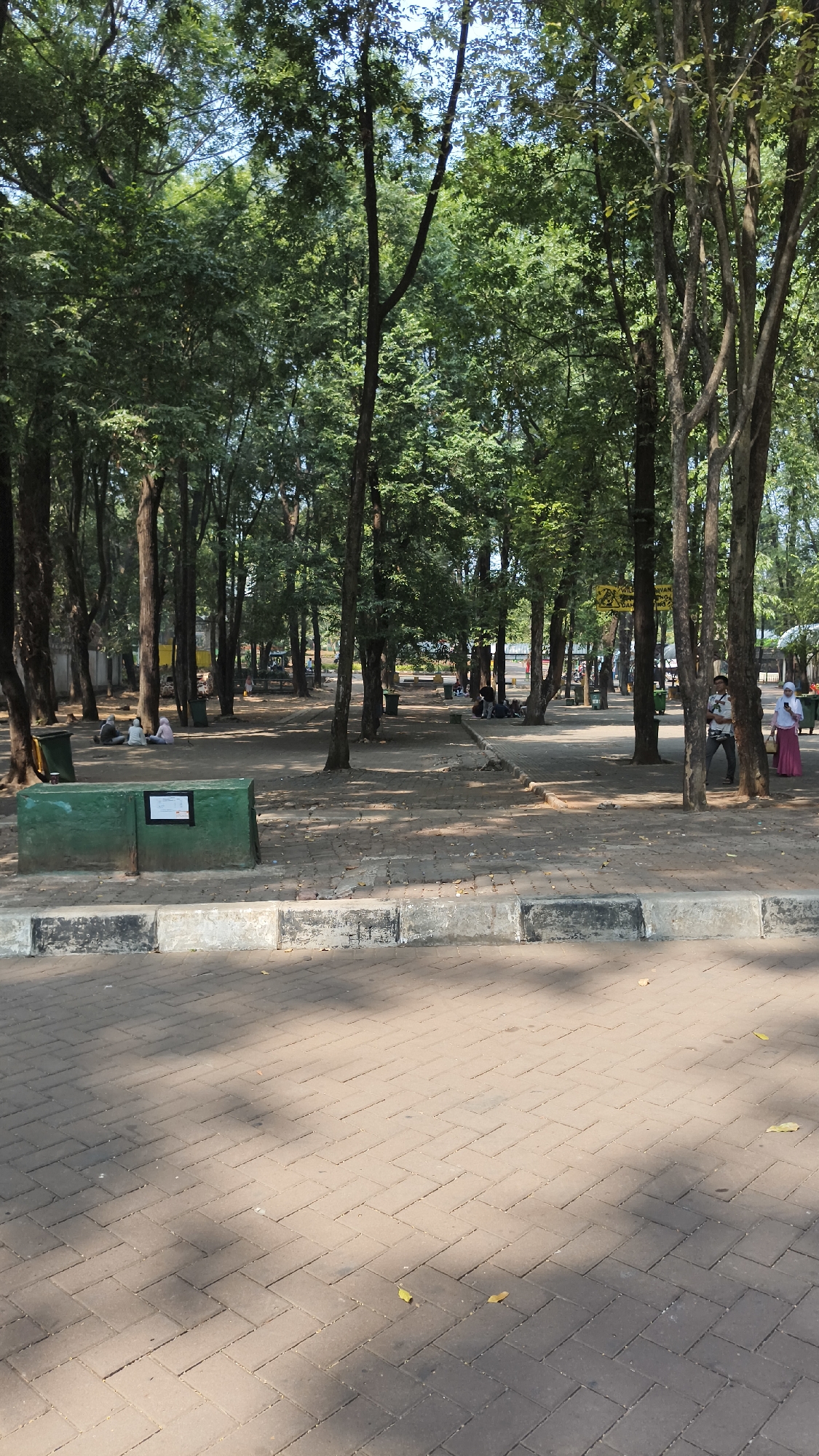 This is a medium-angle outdoor shot taken at Taman literasi Martha Christina Tiahahu in South Jakarta, Indonesia. The image depicts a park setting with a paved walkway in the foreground and a tree-lined area in the background. Several people are scattered throughout the park, some sitting on the ground or on benches, and others walking.
In the foreground, the walkway is paved with light brown interlocking brick pavers, with a concrete curb separating it from a patch of dirt and stone ground. The curb is painted with alternating black and white stripes.
In the middle ground, a green rectangular planter box is present, with a sign on its side. Beyond the planter, the ground is covered with dirt and small stones, with scattered leaves. Several people are seated in small groups on the ground in this area, appearing to be relaxing or socializing. Two green trash receptacles are visible, one to the left of the planter box and another further into the park.
The background is dominated by a dense collection of trees with green foliage, creating a shaded canopy over the park. The trees are of varying sizes and heights, with a mix of thick trunks and thinner branches. Sunlight filters through the leaves, casting dappled shadows on the ground.
In the far background, glimpses of buildings and other park features are visible, including what appears to be a yellow sign. A few individuals are visible in the distance, including a man and two women wearing headscarves. The sky is visible above the trees, appearing bright and clear. The overall impression is of a peaceful, well-maintained urban park.