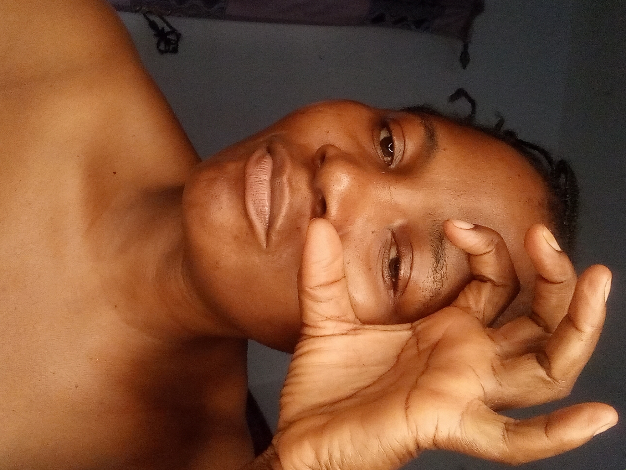A medium shot shows the upper body of a Black woman with dark skin. She is looking directly at the camera with a slight smile. Her right hand is raised, with her thumb and index finger forming a circle around her right eye, obscuring the pupil. Her hair is styled in dark braids that are pulled back. Her left eye is partially visible. The background is a dark, textured wall. The lighting suggests the image was taken indoors, possibly at night or in a dimly lit room. The woman is wearing no visible clothing above the shoulders. The scene is a close-up portrait, focusing on the woman's face and hand gesture.
