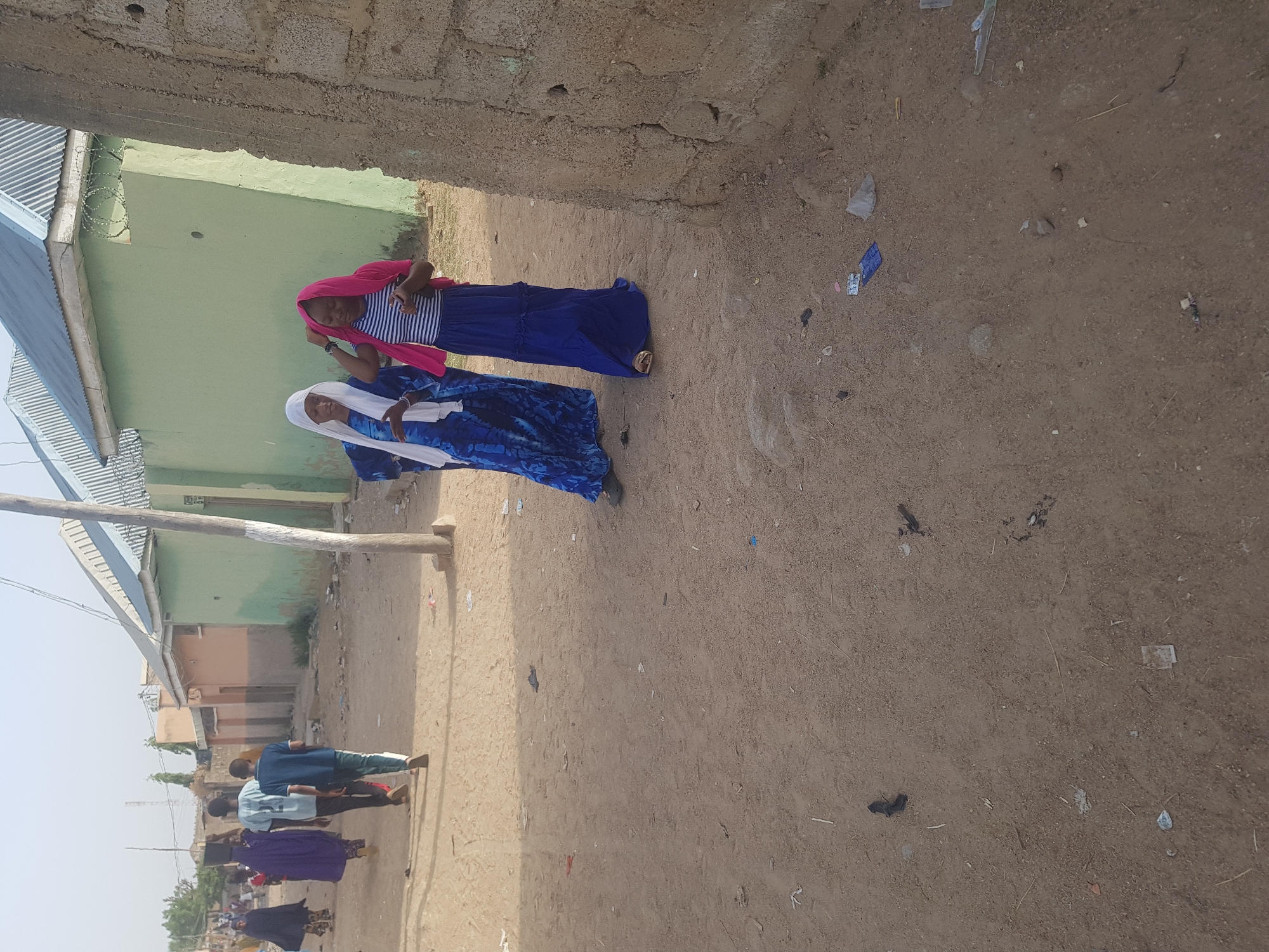 In Dabawa, Nigeria, two women walk along a dirt street enclosed by building walls. One wears a white headscarf and a blue patterned dress. The other wears a pink head covering, a striped shirt, and a blue skirt. Other people walk further down the street in the distance. Trash litters the dirt street. Buildings with corrugated metal roofs are visible.
