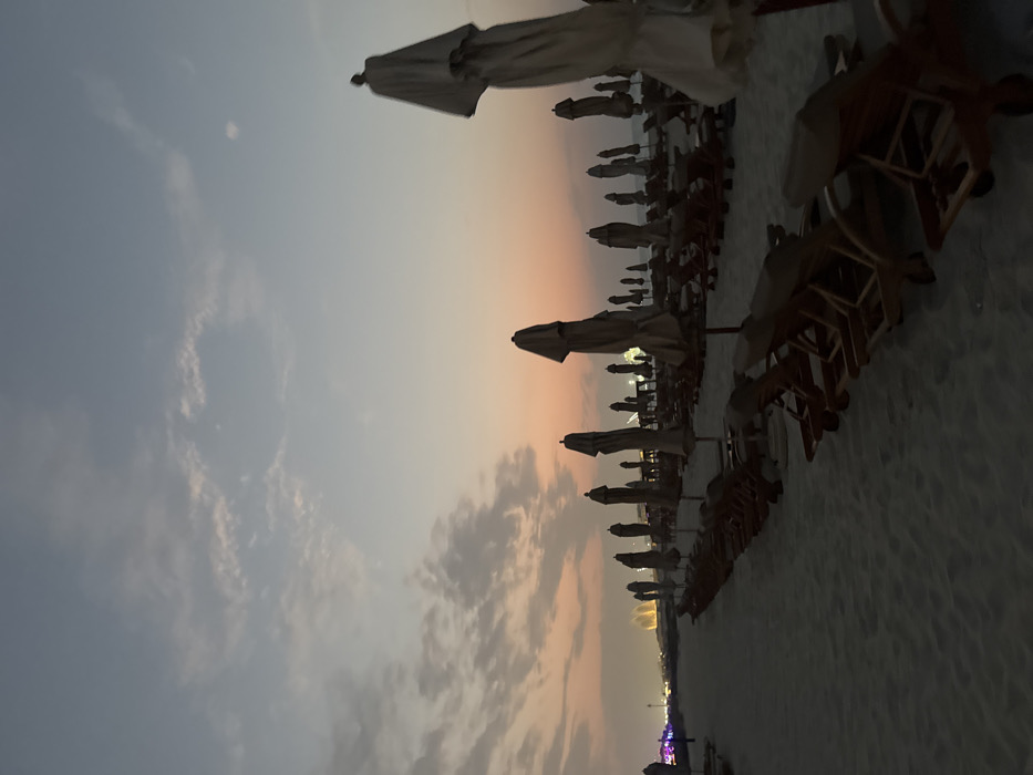 The image displays an unoccupied beach scene at dusk or sunset in Abu Dhabi, United Arab Emirates, with distant structures resembling the Etihad Towers visible on the left horizon, indicating a view towards the Corniche area. The foreground and midground are dominated by numerous rows of closed, light-colored beach umbrellas and associated empty wooden beach loungers, uniformly arranged on light-colored sand. A sandy path is visible on the left side of the frame. No individuals are present. The sky transitions from a soft orange and pink hue near the horizon to a lighter blue-grey overhead, featuring scattered, indistinct cloud formations. Faint city lights are visible along the distant left horizon.