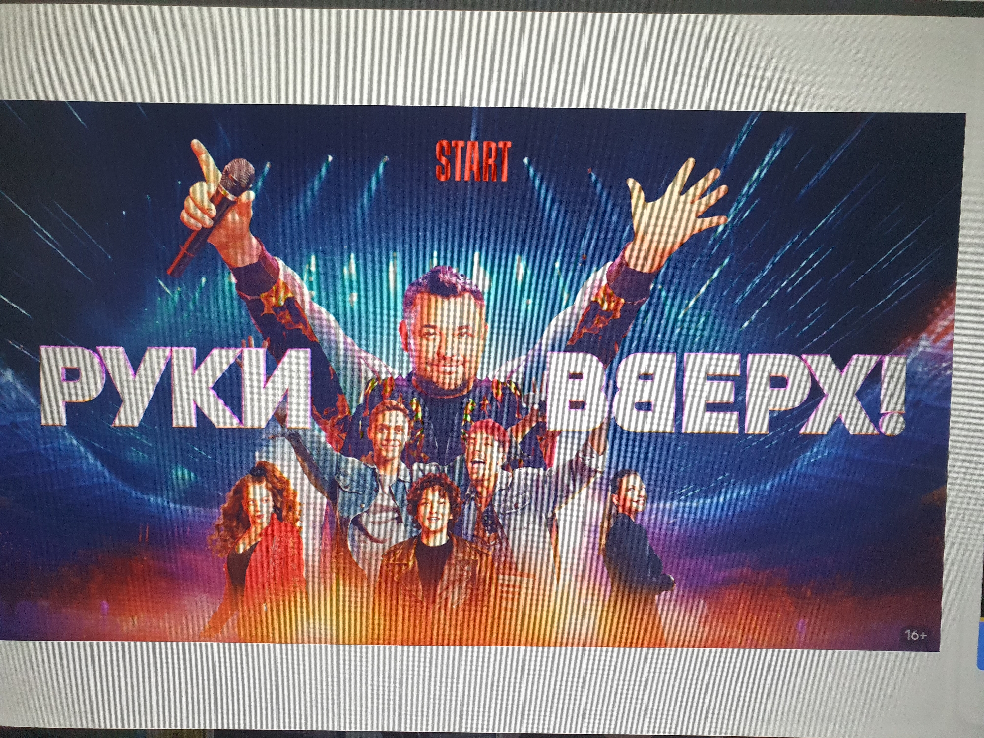 The image is a promotional poster for a concert or event. It features a group of people on a stage with a vibrant, illuminated background. In the foreground, a man holding a microphone is prominent, with his hand raised. To his left and right are other individuals, also seemingly performers, with various expressions and gestures. Large, stylized text that appears to be in Russian, "РУКИ ВВЕРХ!", dominates the lower half of the poster. The word "START" is displayed in red text near the top. A "16+" age rating is visible in the bottom right corner. The image does not contain any information that would indicate a specific location in Tashkent, Uzbekistan, or any other city.