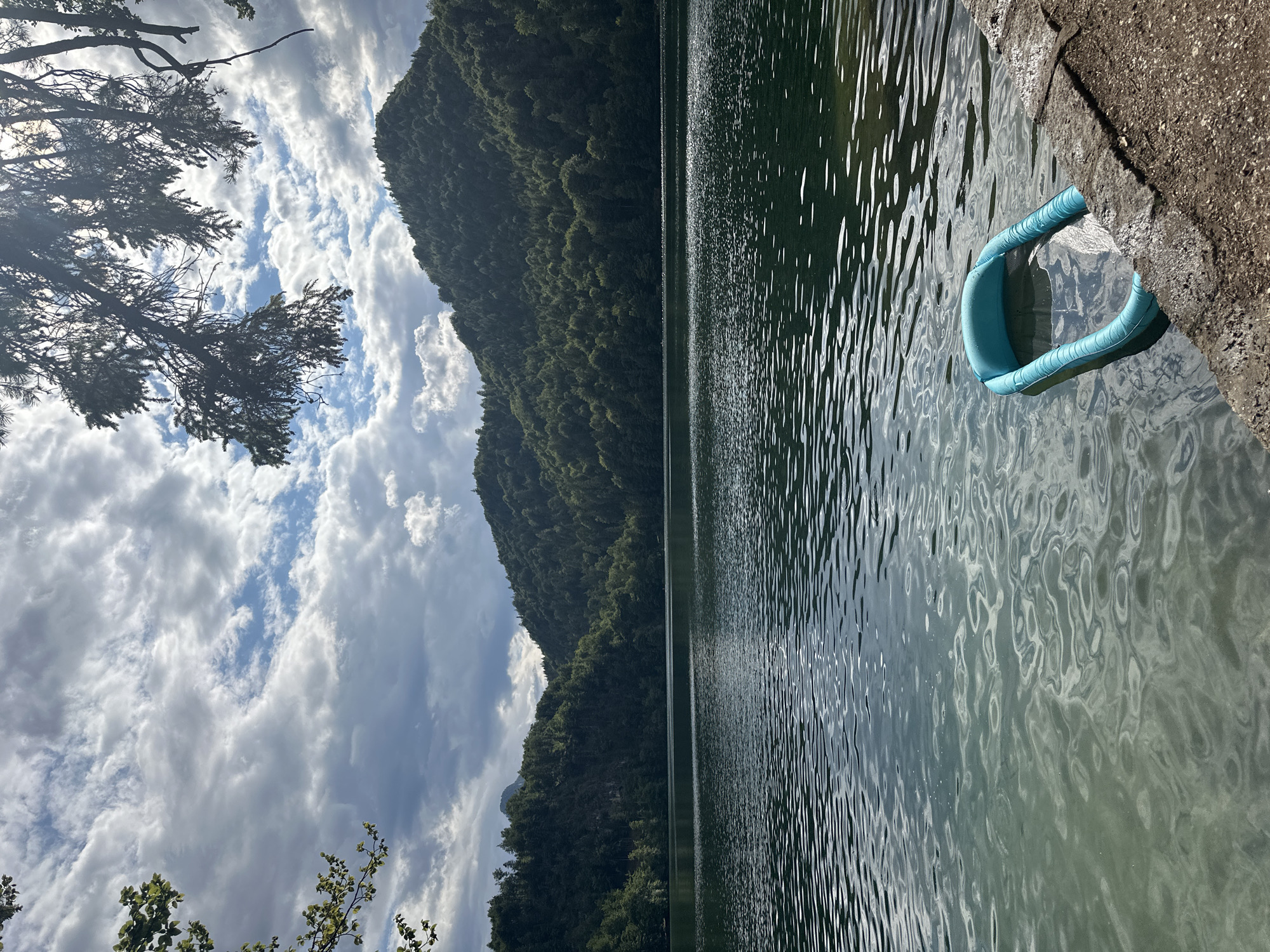 The image shows a tranquil lake scene with a backdrop of lush green mountains under a partly cloudy sky. In the foreground, a small, inflatable blue pool floats on the calm water near the rocky shore. The lake's surface is rippled, reflecting the sky and surrounding foliage. The scene evokes a sense of peace and natural beauty, characteristic of the Bavarian Alps, which surround Kiefersfelden, Germany. The location is likely a popular spot for recreation and enjoying the outdoors.