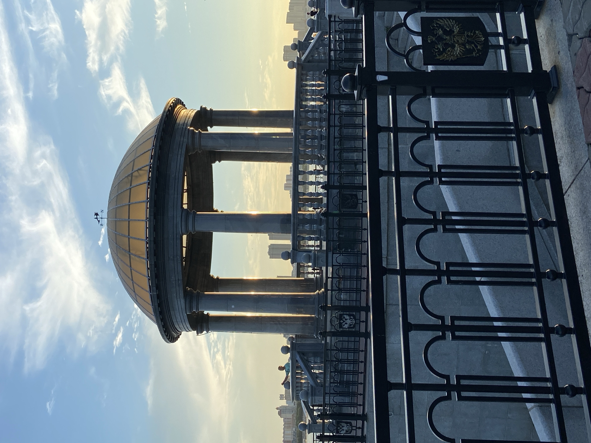 The image depicts a monumental gazebo with a golden dome, supported by large stone columns, situated on a waterfront promenade in Blagoveshchensk, Russia. The gazebo is encircled by a stone balustrade and a black ornate metal fence, which also frames a staircase leading down. In the background, the Amur River is visible, with a city skyline of buildings and high-rises on the opposite bank, suggesting the border with China. The sky is a vibrant blue with scattered wispy clouds, and the setting sun casts a warm golden light on the gazebo and the surrounding architecture, creating long shadows. A weathervane is visible atop the dome. Two individuals are present: one person stands near the balustrade within the gazebo area, and another is seen further in the background along the promenade. The scene conveys a sense of public space and architectural grandeur in an urban waterfront setting during sunset.