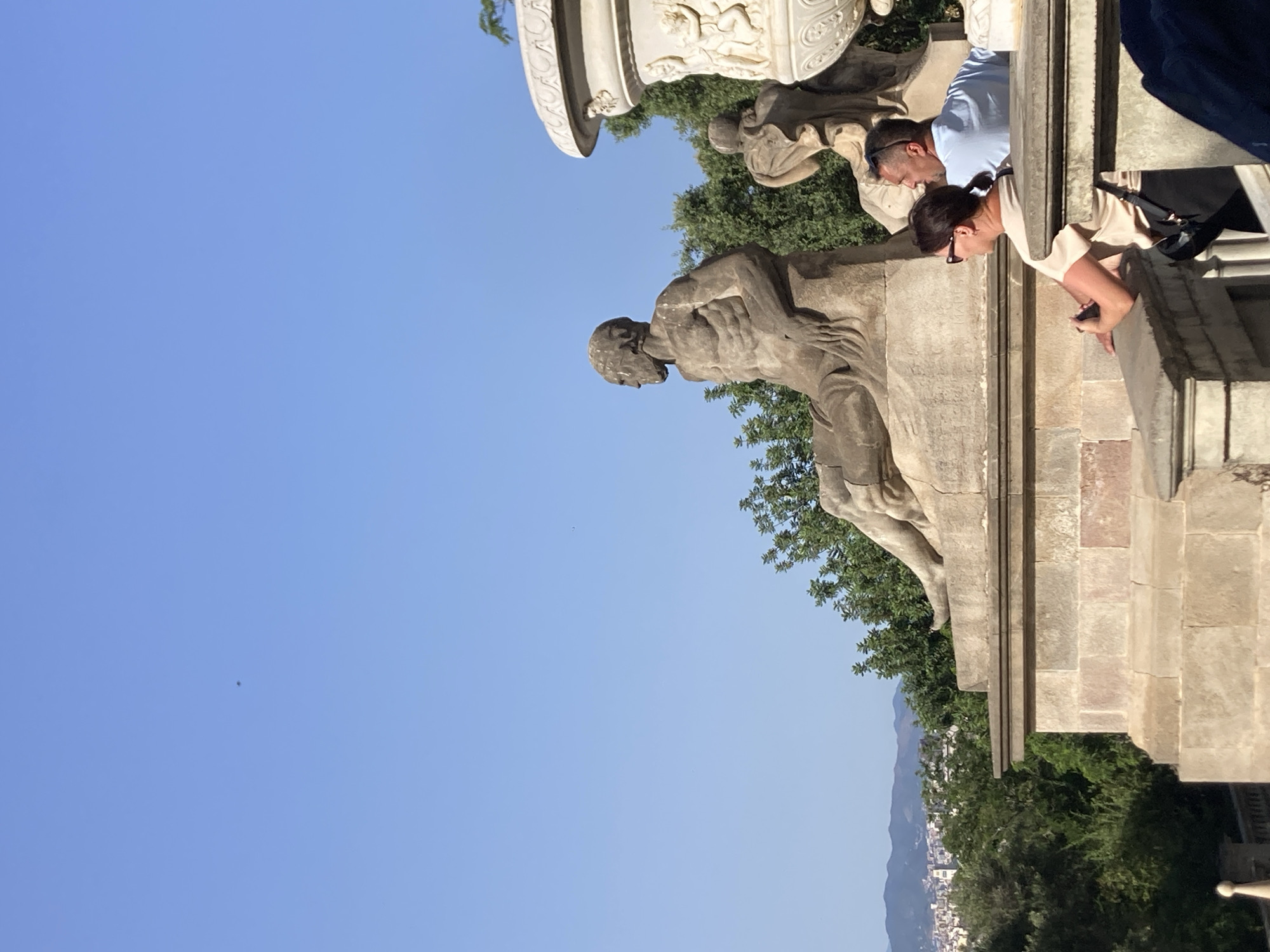The image depicts a sunny outdoor scene with a clear blue sky. In the foreground, two people, a woman and a man, are seated on a stone structure, looking to their right. The woman is wearing a beige top and sunglasses, with her hair in a ponytail. The man is wearing a light blue t-shirt and sunglasses perched on his head.
Behind them, a large stone sculpture of a reclining nude male figure is prominent. To the right of this sculpture is a decorative stone urn. In the background, lush green trees are visible, and beyond that, a city skyline with buildings and mountains can be seen, suggesting this location is elevated within the city of Barcelona, Spain. The overall atmosphere is relaxed, possibly in a park or a viewpoint area.