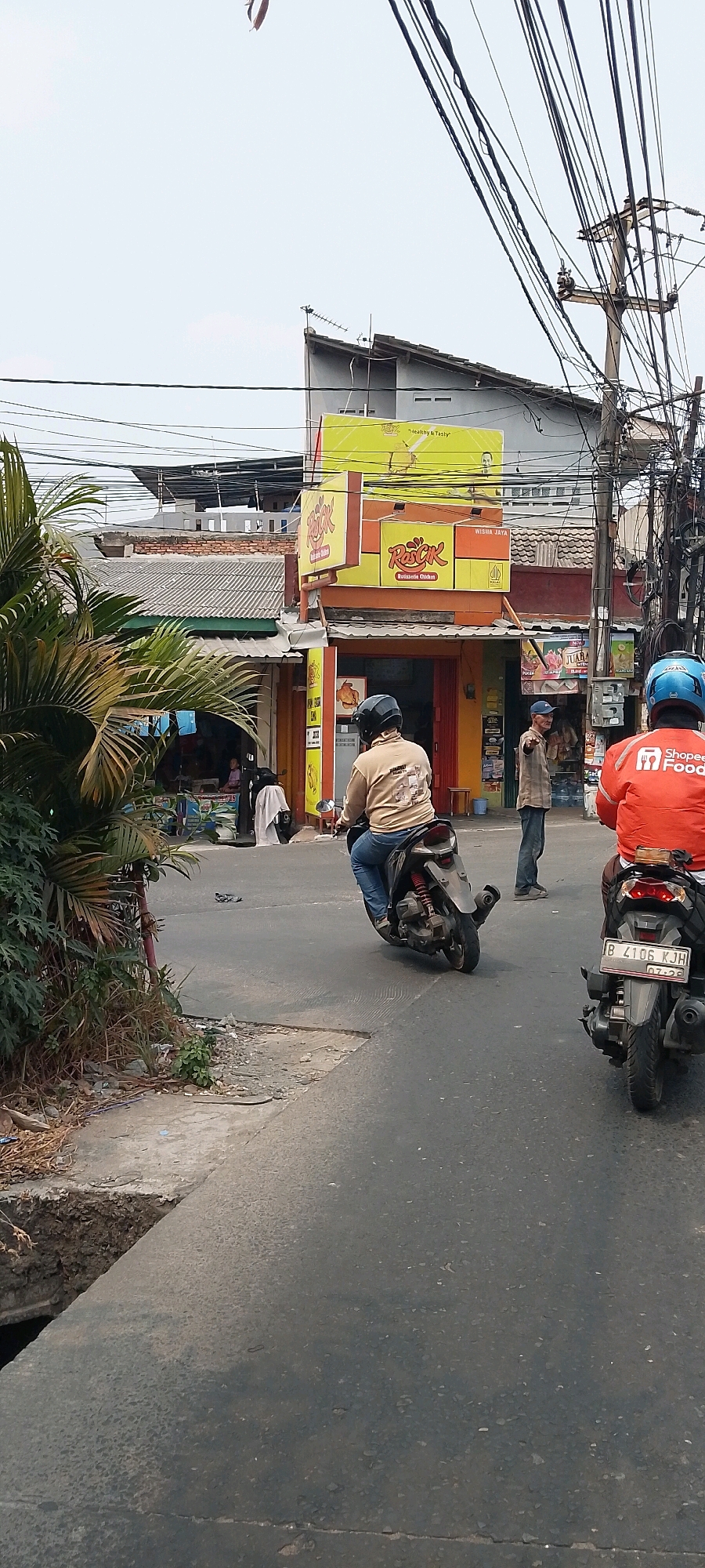 The image shows a street scene in Bekasi, Indonesia. Two individuals on motorcycles are the primary subjects. The person in the foreground is wearing an orange jacket with a Shopee Food logo, indicating they are likely a delivery driver. They are riding a black motorcycle. Behind them, another person in a tan jacket and blue jeans is also on a motorcycle, which is dark gray with red rear suspension. A man in a plaid shirt and blue cap is standing near a building with a yellow sign that reads "Rasck Rotisserie Chicken." The building appears to be a food establishment. There are power lines overhead, suggesting an urban environment. A palm tree is visible on the left side of the image. The road surface is asphalt with visible cracks.