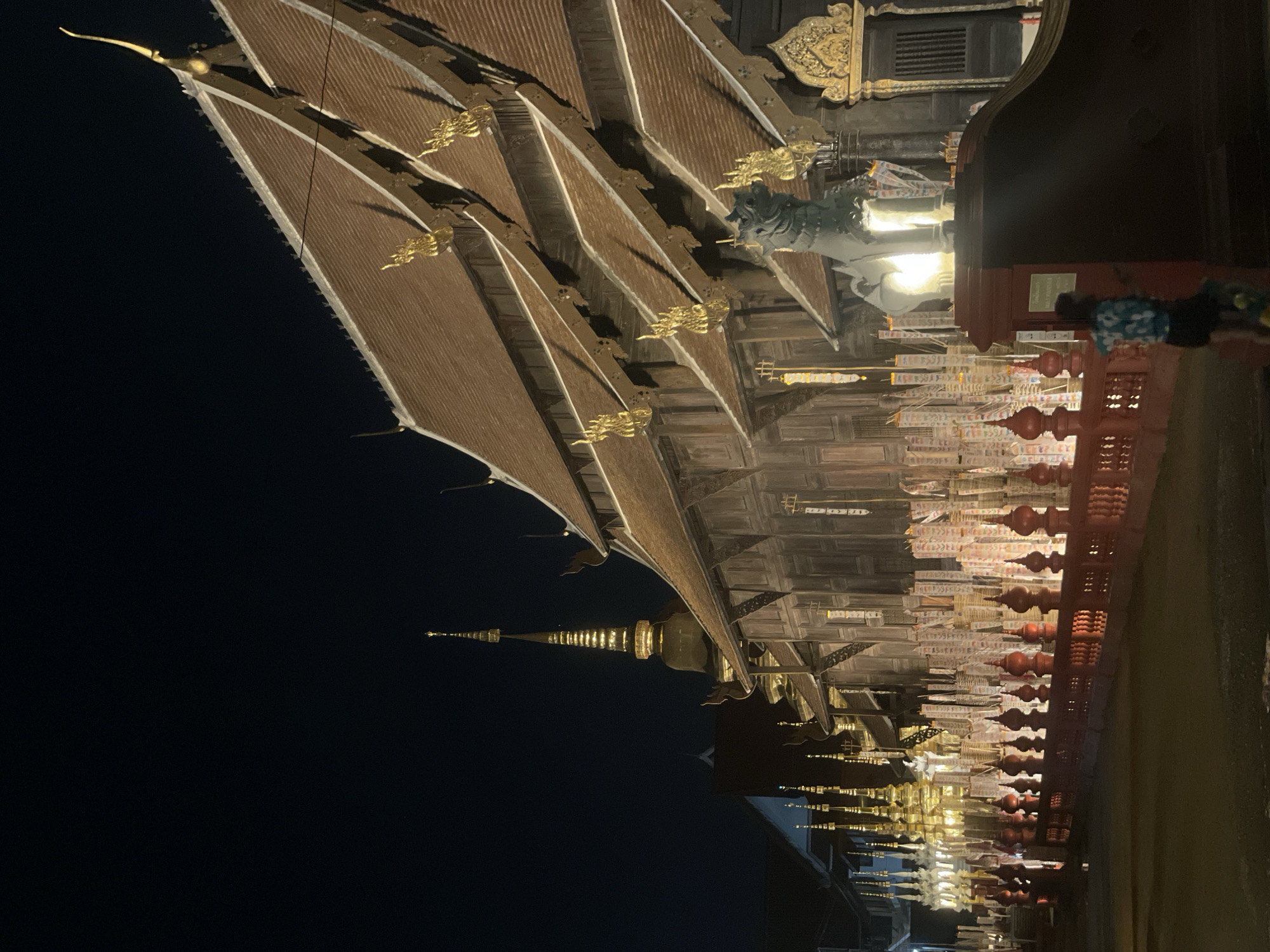 The image depicts a low-angle view of a temple in Chiang Mai City Municipality, Thailand, at night. The temple features multiple layers of roofs with ornate golden decorations. A golden spire is visible in the distance. A row of lanterns adorns the temple's facade.
