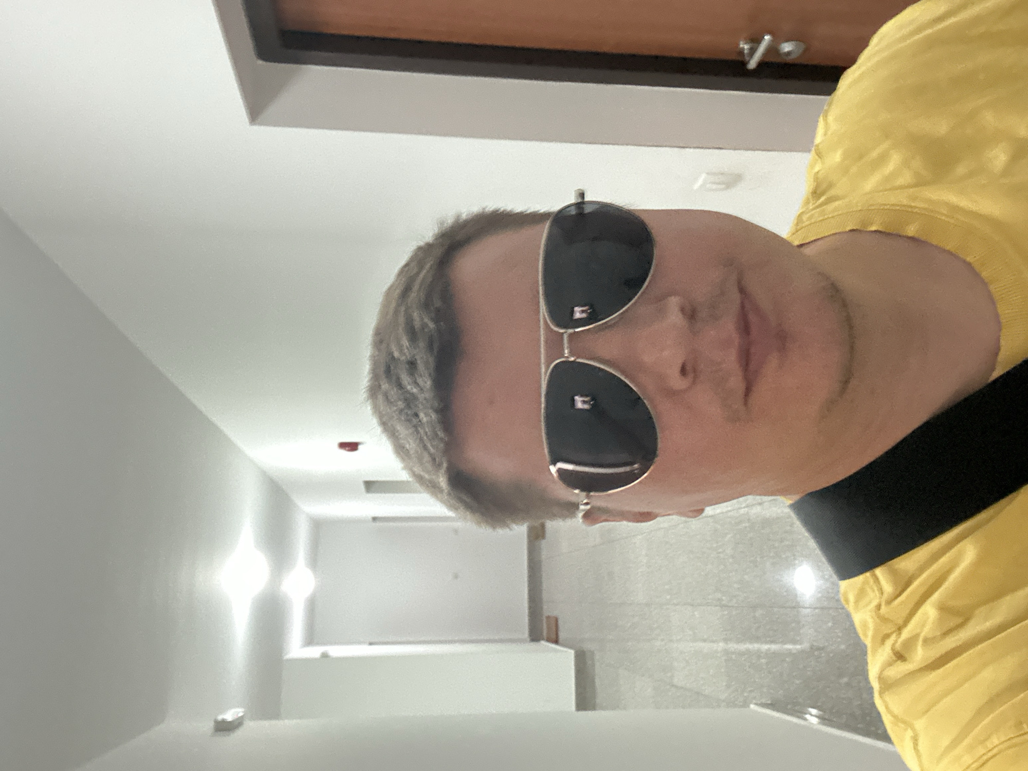 A man wearing sunglasses and a yellow t-shirt takes a selfie in what appears to be a hotel corridor. The man is facing the camera, and his head is tilted. The sunglasses are aviator-style with black lenses and a silver frame. The yellow t-shirt has a black stripe across the chest. The corridor is brightly lit with white walls and a tiled floor. There are lights on the ceiling, and a door is visible at the end of the corridor. The time of day is not specified, but the lighting suggests it could be daytime. The overall mood of the image is casual and candid. There are no visible text elements in the image. The location context provided indicates Budva, Montenegro.