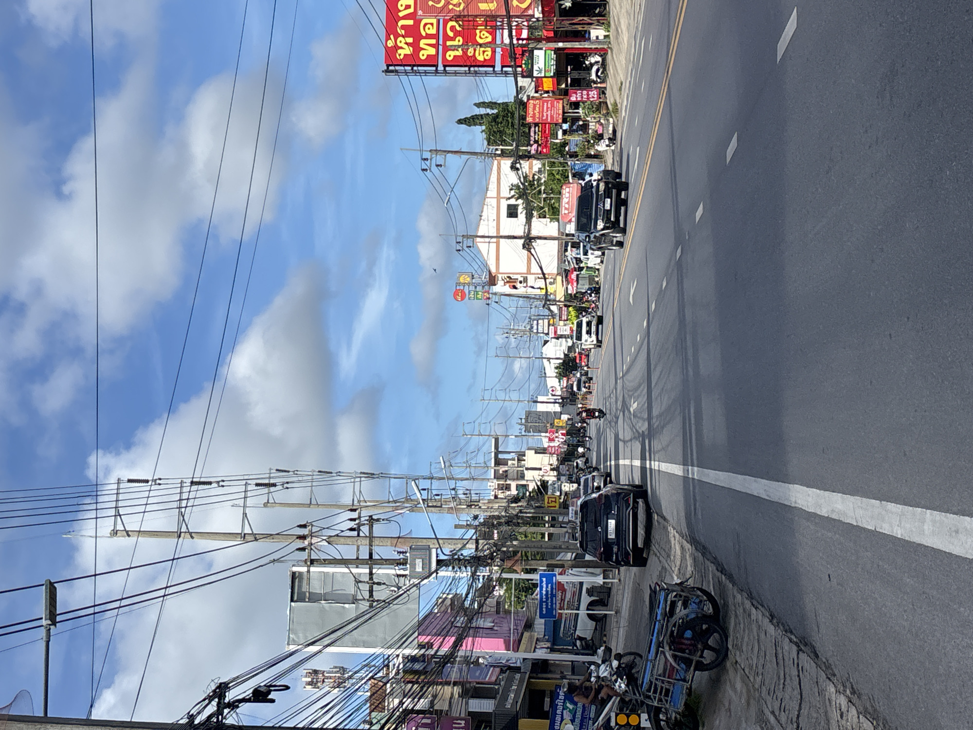 This is a vertical, eye-level, medium-range shot of a street scene in Chalong, Thailand, on a partly cloudy day. The street is asphalt with yellow and white lane markings. On the left side of the road, there is a sidewalk with a parked motorcycle with a sidecar, a utility pole with numerous wires, and several shops with colorful signs. On the right side of the road, there are more shops and buildings, also with prominent signs. Several vehicles, including cars and motorcycles, are on the road, some moving and some parked. A few people are visible, including a person on a motorcycle on the left sidewalk and several figures on motorcycles and as passengers on the right side of the road. The sky is blue with scattered white clouds. The overall impression is a busy, commercial street in a Southeast Asian city.