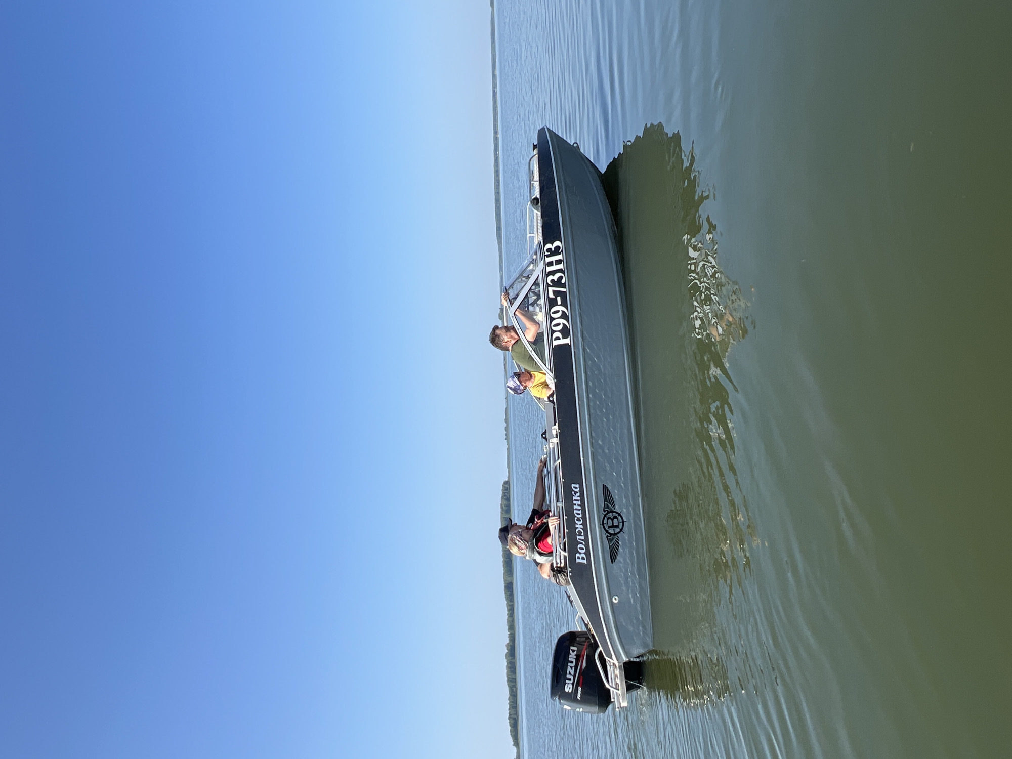 The image shows a gray motorboat with "P99-73H3" and "Волжанка" written on its side, moving across a body of calm water under a clear blue sky. Three people are visible in the boat: two adults and a child. One adult is steering, while the other two are seated. A Suzuki outboard motor is attached to the stern of the boat. The environment appears to be a large lake or river, with a distant treeline visible on the horizon. The water is green and reflects the boat and sky. This scene is likely taking place near Novopichugovo, Russia, a village located on the shore of Lake Shartash, which fits the description of a large body of water with a treeline.