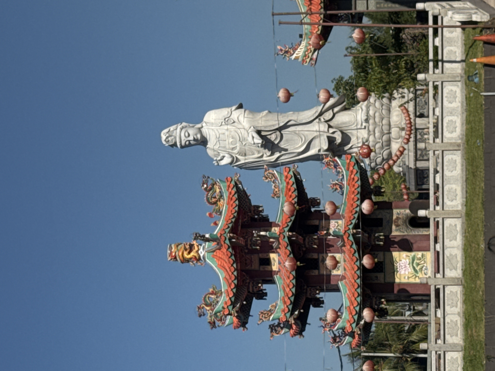 The image shows a marble statue of a female figure, possibly a deity, standing atop an elaborately decorated, multi-tiered pagoda structure. The pagoda features red, green, and brown colors with dragon motifs and numerous red lanterns hanging from its eaves. The background is a clear blue sky. The scene is likely located in Denpasar, Indonesia, given the architectural style and cultural elements present.