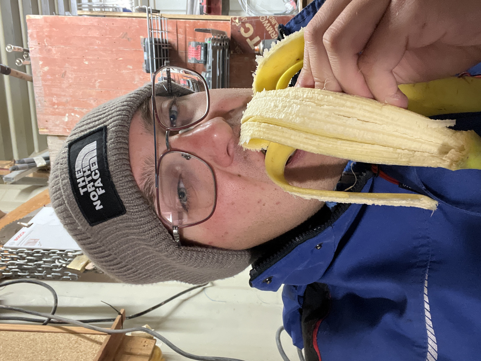 A close-up, tilted shot shows a person with glasses and a gray beanie eating a peeled banana. The beanie has a "The North Face" logo. The person is wearing a blue jacket. The background includes a red wooden structure with metal tools, chains, and wiring. The location in Varna, Bulgaria, is not discernible from the visual information provided.
