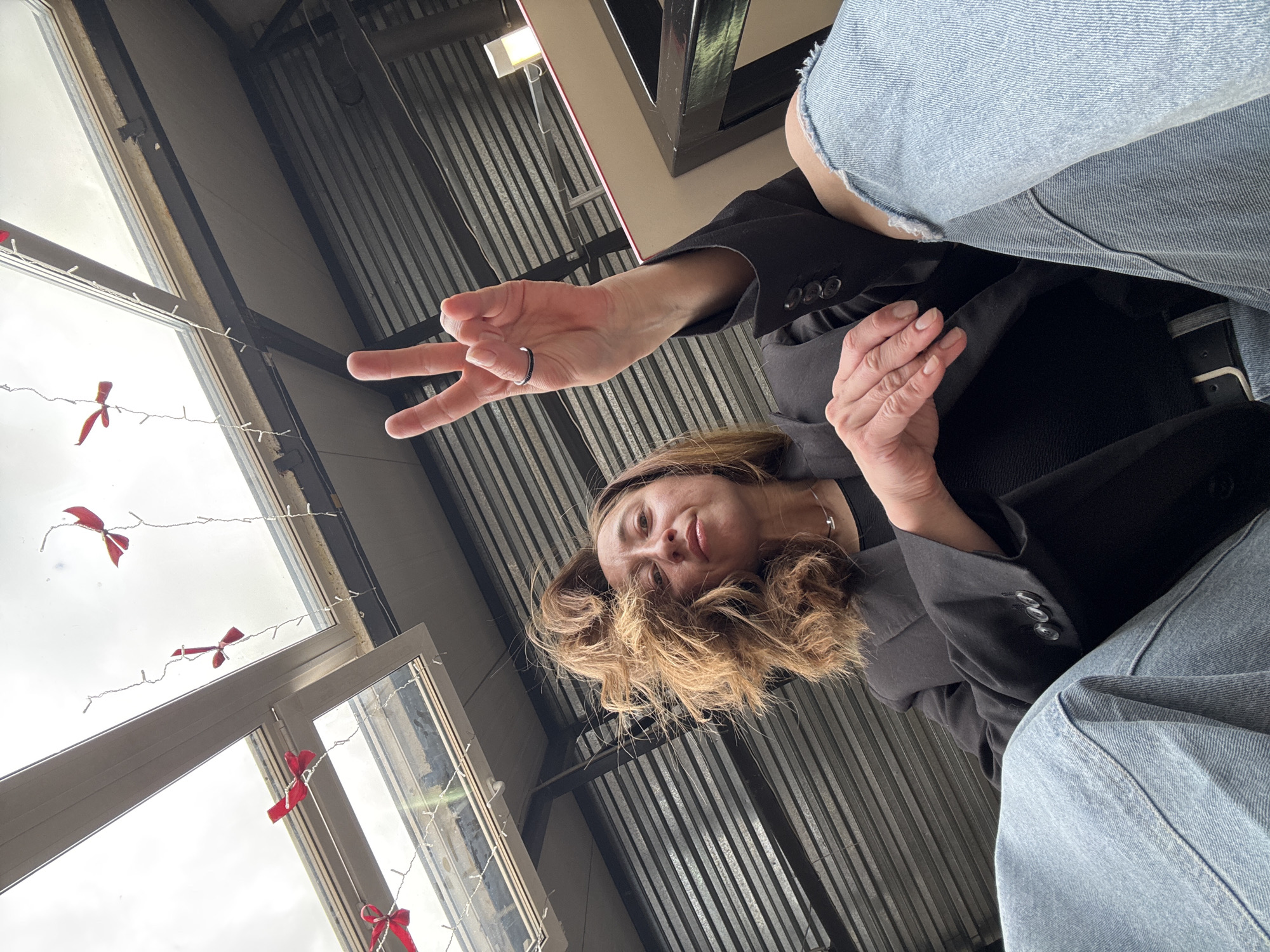 The image captures a woman from a low, upward-looking perspective within an interior space, likely in Chaikovsky, Russia. The woman has light brown, wavy hair and is dressed in a black blazer over a darker top, paired with light-wash blue jeans. She is looking down towards the camera with a neutral, slightly playful expression. Her right hand is raised, forming a peace sign, with a ring visible on her ring finger, while her left hand rests casually on her lap.
The background to the left features a large window or skylight, revealing a bright, overcast sky. Fairy lights decorated with small red bows are strung across the window panes, adding a festive or decorative element to the setting. To the right and in the upper portion of the frame, a corrugated metal structure, possibly a ceiling or wall, suggests an industrial or warehouse-like interior. The overall lighting is bright and even, consistent with daylight on an overcast day. There is no visible text in the image.