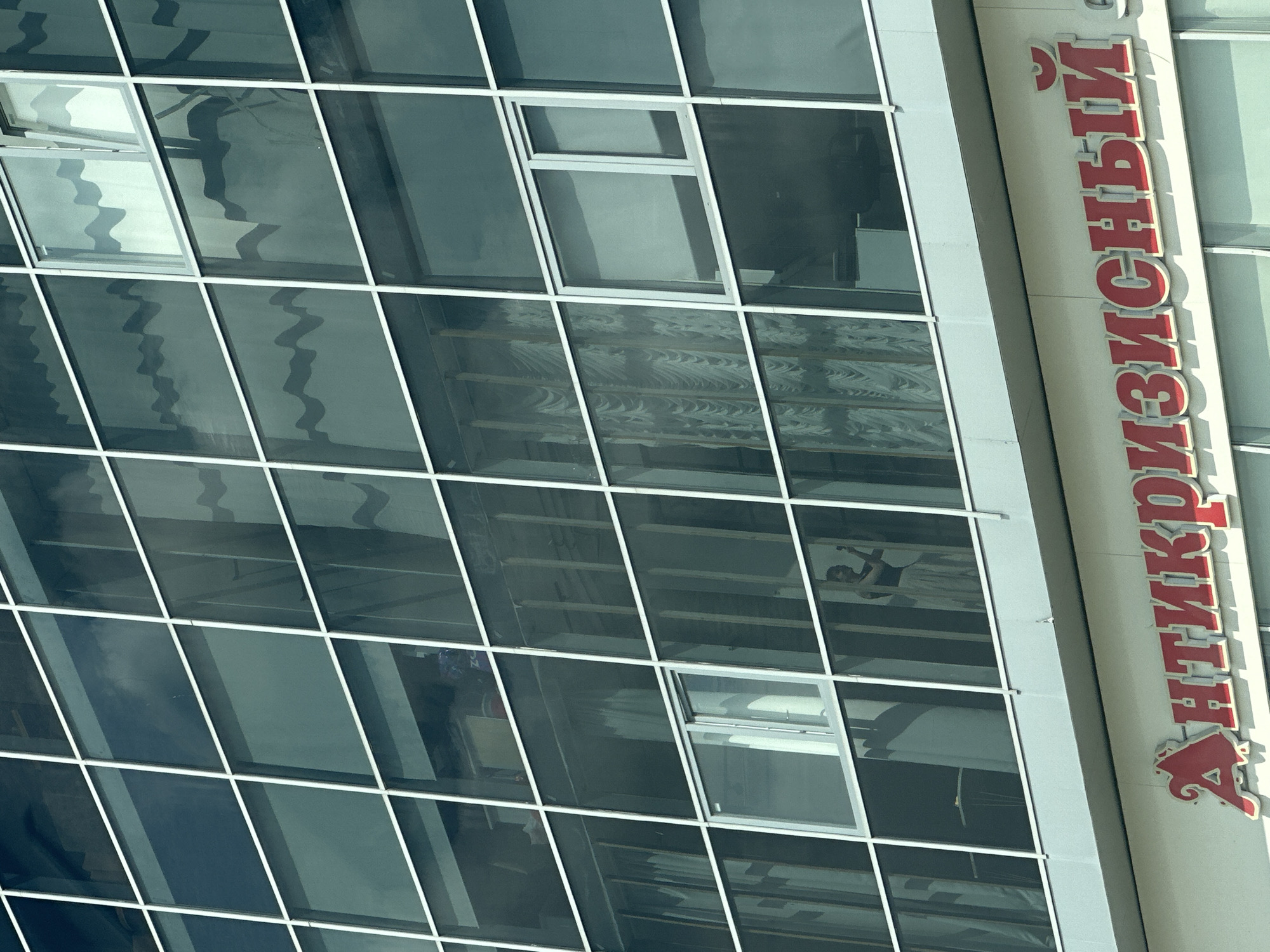 The image shows a close-up of a modern building with a glass facade. The building has a grid of white frames, dividing the glass into numerous windows. Reflections of clouds and wavy patterns are visible on the glass. In one of the windows, a person appears to be standing and looking at their phone. Below the glass facade, a sign with red Cyrillic lettering is visible. The sign reads "АНТИКРИЗИСНЫЙ" which translates to "Anticrisis". This building is located in Magnitogorsk, Russia.