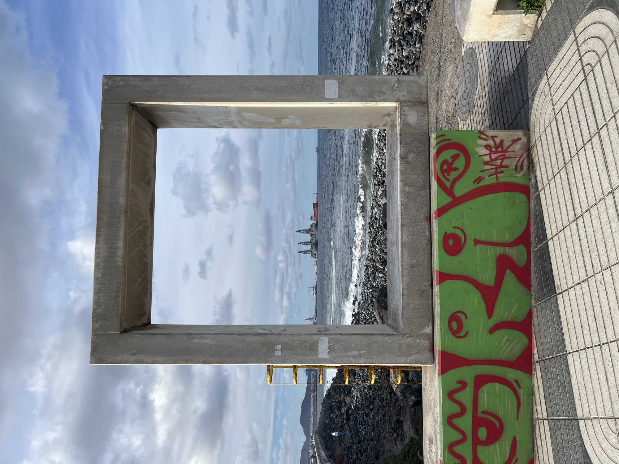 The image shows a view of the ocean framed by a concrete structure in Las Palmas, Spain. The ocean is visible through the square opening of the structure, with a rocky beach in the foreground. There are buildings and ships visible in the distance, further out in the ocean. To the right of the concrete frame, there's a painted or graffitied concrete support. The ground is a paved walkway with a gray and white geometric pattern. In the lower part of the view, below the ocean horizon, there is a rocky shoreline. The sky is overcast with clouds, with a portion visible above the concrete structure.