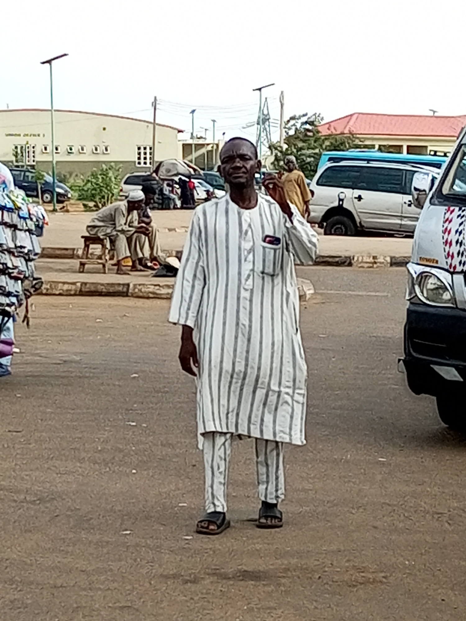 In the city of Gombe, Nigeria, a man stands in the foreground, facing the camera, wearing a white and grey striped traditional outfit and dark sandals. He has his right hand raised to his face. Behind him, a bus with a white and black exterior is parked on the right. To the left of the man, another individual is seated on a wooden stool, wearing a light-colored traditional outfit and a head covering. Several parked vehicles, including vans and a blue bus, are visible in the background, along with a building and streetlights. A market stall with clothing items is situated on the far left of the image.