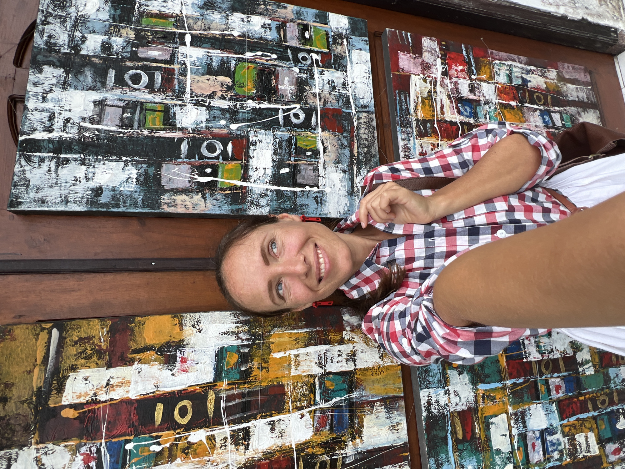 A woman stands in front of four abstract paintings. She is facing the viewer and smiling. She is wearing a red and blue checkered shirt and a white top. The paintings are colorful and feature geometric shapes and lines. The top left painting features a black background with white accents. The top right painting features a red background with white accents. The bottom left painting features a yellow background with white accents. The bottom right painting features a red background with white accents. The paintings are displayed on a wooden surface, and the scene appears to be in the Ubud District of Indonesia.