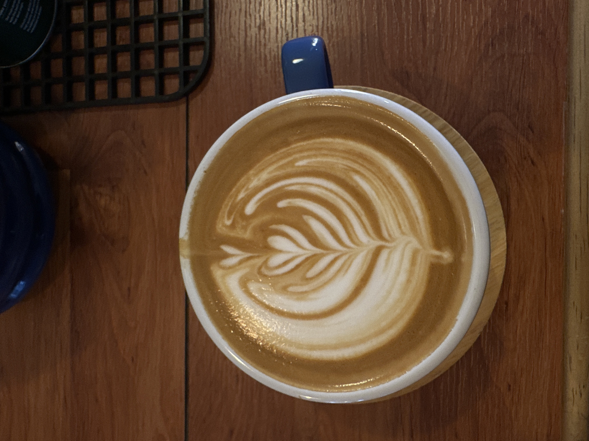 The image shows a close-up, overhead view of a cup of latte art on a wooden surface. The coffee has a creamy brown color with intricate white foam art resembling a fern leaf. A small, blue spoon handle is visible, sticking out of the top of the cup. To the left of the cup, part of a dark blue bowl or cup is visible. In the upper left corner, a portion of a black, grid-like object, possibly a placemat or coaster, is also present. The wooden surface appears to be a table with a visible grain. The lighting suggests an indoor setting, likely a cafe. No people are visible in the frame. There is no discernible time of day, weather, or specific location cues beyond the general setting. No text is visible in the image.