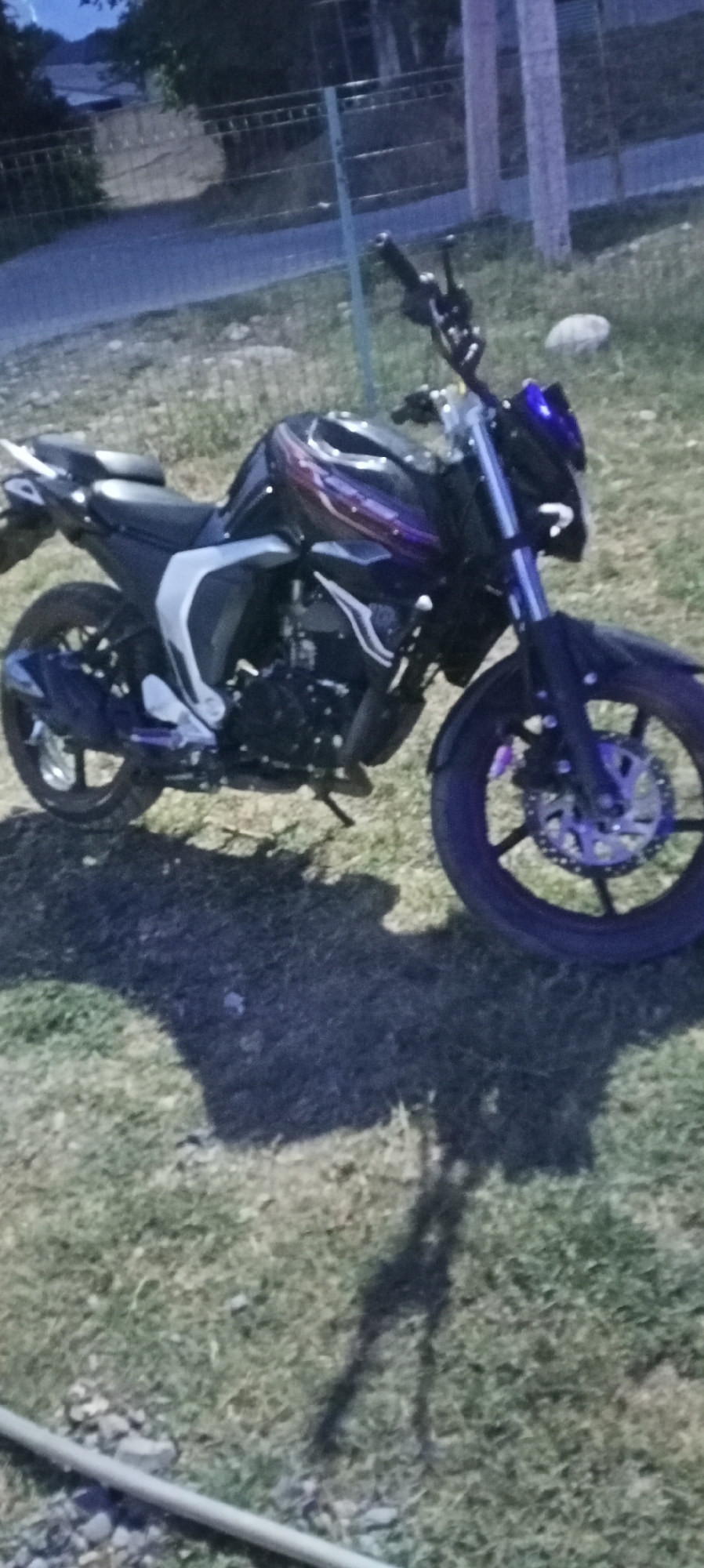 The image shows a parked motorcycle on grassy ground, with a paved area and a fence visible in the background. The motorcycle is dark colored with purple and white accents. The front wheel and handlebars are visible on the right side of the image, and the rear wheel is visible on the left. There is a shadow cast by the motorcycle on the ground. The environment appears to be outdoors, possibly a street or park, with some vegetation and a fence. The image quality is poor, with low resolution and motion blur, making it difficult to discern fine details. There are no people or explicit signs indicating the location in Sokuluk, Kyrgyzstan.