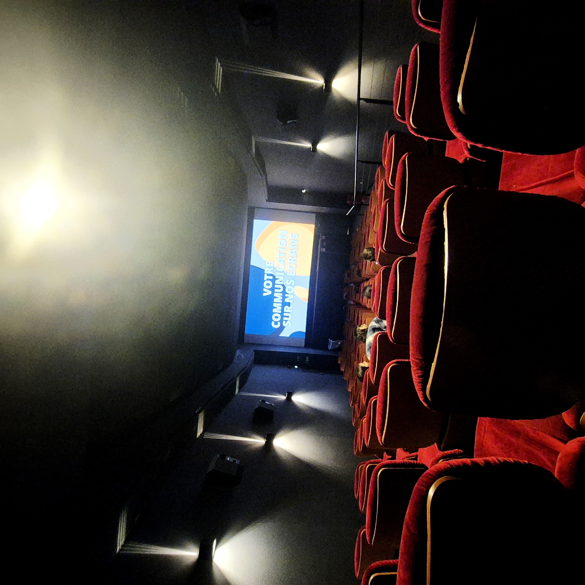 An interior shot of a darkened cinema in Strasbourg, France. Rows of red velvet seats dominate the right side of the image, leading towards a bright screen at the far end. The screen displays the text "VOTRE COMMUNICATION SUR NOS ÉCRANS" in white letters against a blue and orange background. A few people can be seen seated in the rows of chairs, looking towards the screen. Wall-mounted spotlights illuminate the aisle, casting beams of light. The left side of the image is predominantly dark.