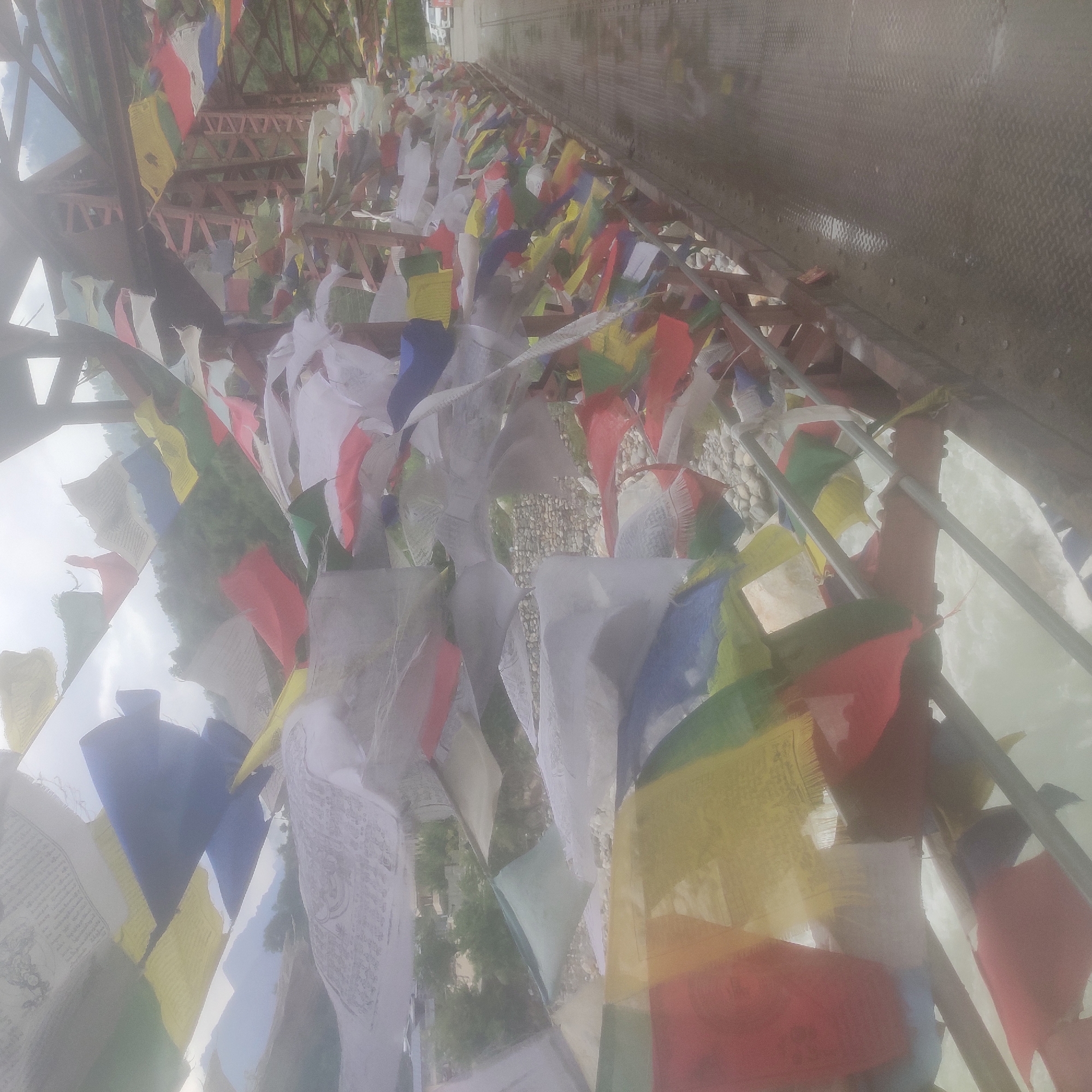 The image shows a bridge decorated with numerous colorful prayer flags, characteristic of Buddhist traditions. The bridge appears to be an infrastructure element crossing a body of water, possibly a river, as indicated by the turbulent white water visible beneath. The flags are of various colors including blue, yellow, red, green, and white, and are stretched across the railing of the bridge, fluttering in the wind. Some flags have visible text or patterns on them. The bridge structure itself is made of metal with a reddish-brown hue, with visible beams and supports. The surrounding environment appears to be mountainous, with green vegetation and misty hills visible in the background. The ground surface of the bridge walkway is metallic and appears wet, suggesting recent rain or mist. In the far distance, a vehicle can be seen on a road. The overall impression is that of a scenic location, likely in a mountainous region of India, specifically Kalath, which is known for its natural beauty and Buddhist cultural influences.