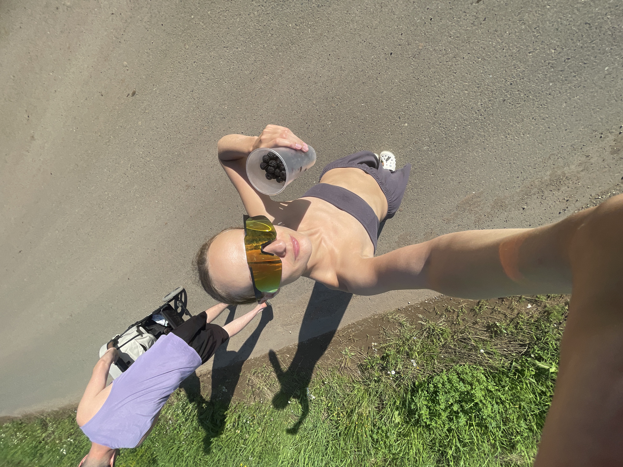 In the city of Podshivalovo, Russia, on a paved road bordered by lush green grass and wild flowers, a woman is captured in a selfie, holding a transparent cup filled with dark berries. She is wearing a grey sports bra and matching sweatpants, with distinctive white crocs on her feet. Her mirrored sunglasses reflect the bright sunlight. Behind her, a man in a lavender tank top and black shorts pushes a light-colored stroller along the road. The scene is illuminated by the sun, casting long shadows on the ground, indicating it is likely late afternoon or early morning. The overall impression is of a casual outdoor activity, possibly a walk or a stroll, with the berries suggesting a potential stop for foraging or a snack.