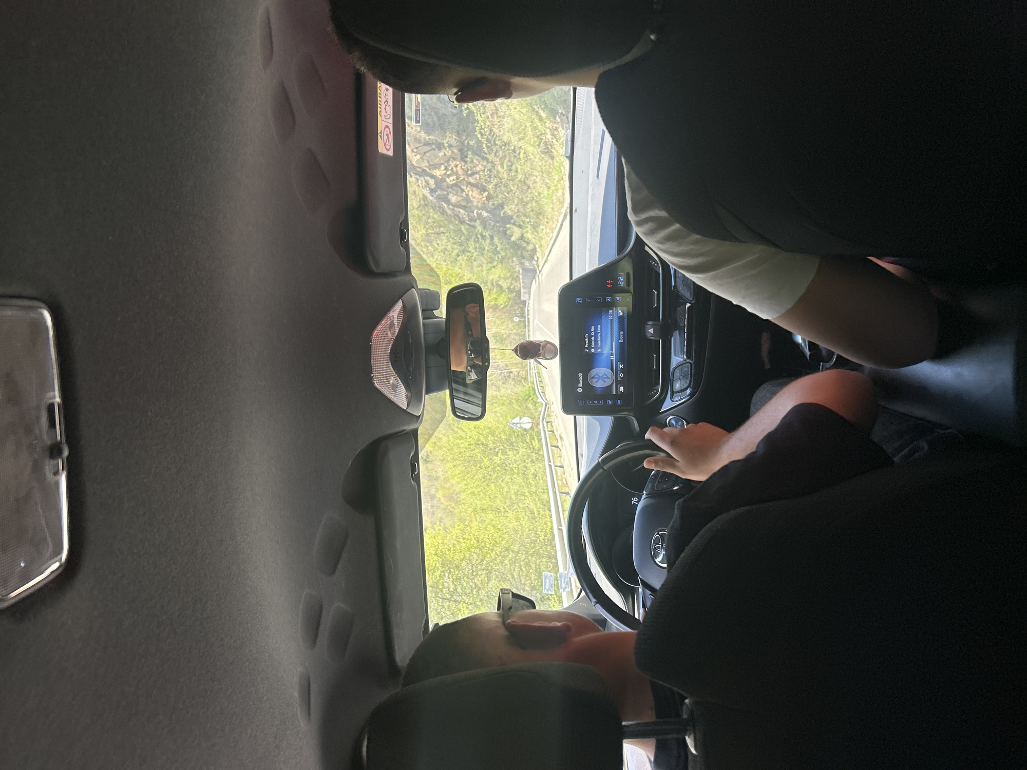 A view from the rear passenger seat of a car captures a daytime journey through Taran, Bulgaria. In the foreground, the head of a person seated behind the driver is partially visible, looking forward. The driver, wearing glasses, has hands on the steering wheel, which bears the Toyota logo and control buttons.
The car's interior is prominent, showing the dashboard and an infotainment system displaying "12:21" as the time and playing music by "Kendrick Lamar" titled "Humble," with a Bluetooth icon visible. A small air freshener hangs from the rearview mirror. The passenger-side sun visor features an "AIRBAG" warning sticker.
Beyond the windshield, a winding road is visible, flanked by lush green hills and trees, suggesting a rural or mountainous landscape. A road barrier or guardrail can be seen along the side of the road. The bright light indicates a clear, sunny day. The scene depicts a tranquil moment of travel.