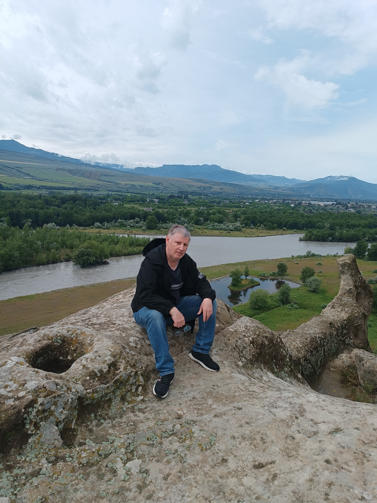A middle-aged man with graying hair sits on a large, rough-textured rock with lichen growth, facing the camera. He wears a black jacket, a t-shirt, blue jeans, and black sneakers. His arms are resting on his knees, and he holds a pair of sunglasses in his right hand. The man is positioned in the foreground of the image, with his legs extending towards the right edge.
Behind him, a vast landscape unfolds. A wide, murky river flows from the left to the right, bordered by a grassy bank. Beyond the river, a dense forest of green trees stretches across the middle ground. In the distance, a town or city with buildings can be seen nestled amongst the trees, and further back, rolling hills and mountains under a cloudy sky complete the scene. The rock formation the man is sitting on appears to be part of a larger cliff or ancient structure, with a noticeable circular indentation or hole in the rock to his left. The environment suggests an outdoor, natural setting, likely a historical or scenic location in Uplistsikhe, Georgia.