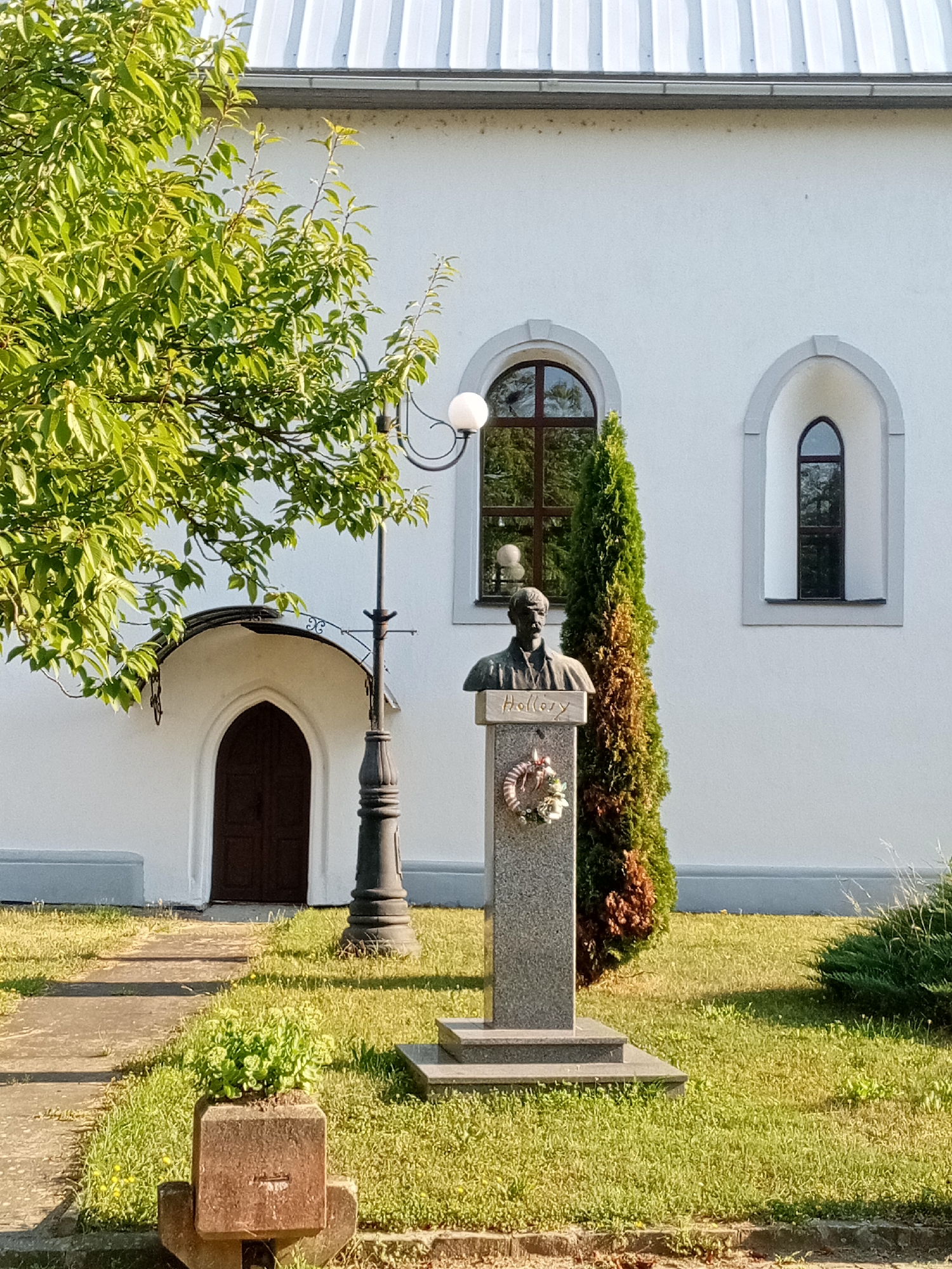 The image shows a bust of a man, identified by the name "Hollery" inscribed on its pedestal, located in front of a white building with arched windows and a metal roof. A cypress tree stands to the right of the bust, and a lamppost is positioned to the left. In the foreground, a concrete planter with green foliage sits on a pathway. The bust and surrounding area are part of a memorial or monument. The scene appears to be outdoors, possibly in a park or on the grounds of a public building. This memorial is located in the city of Tiachiv, Ukraine.
