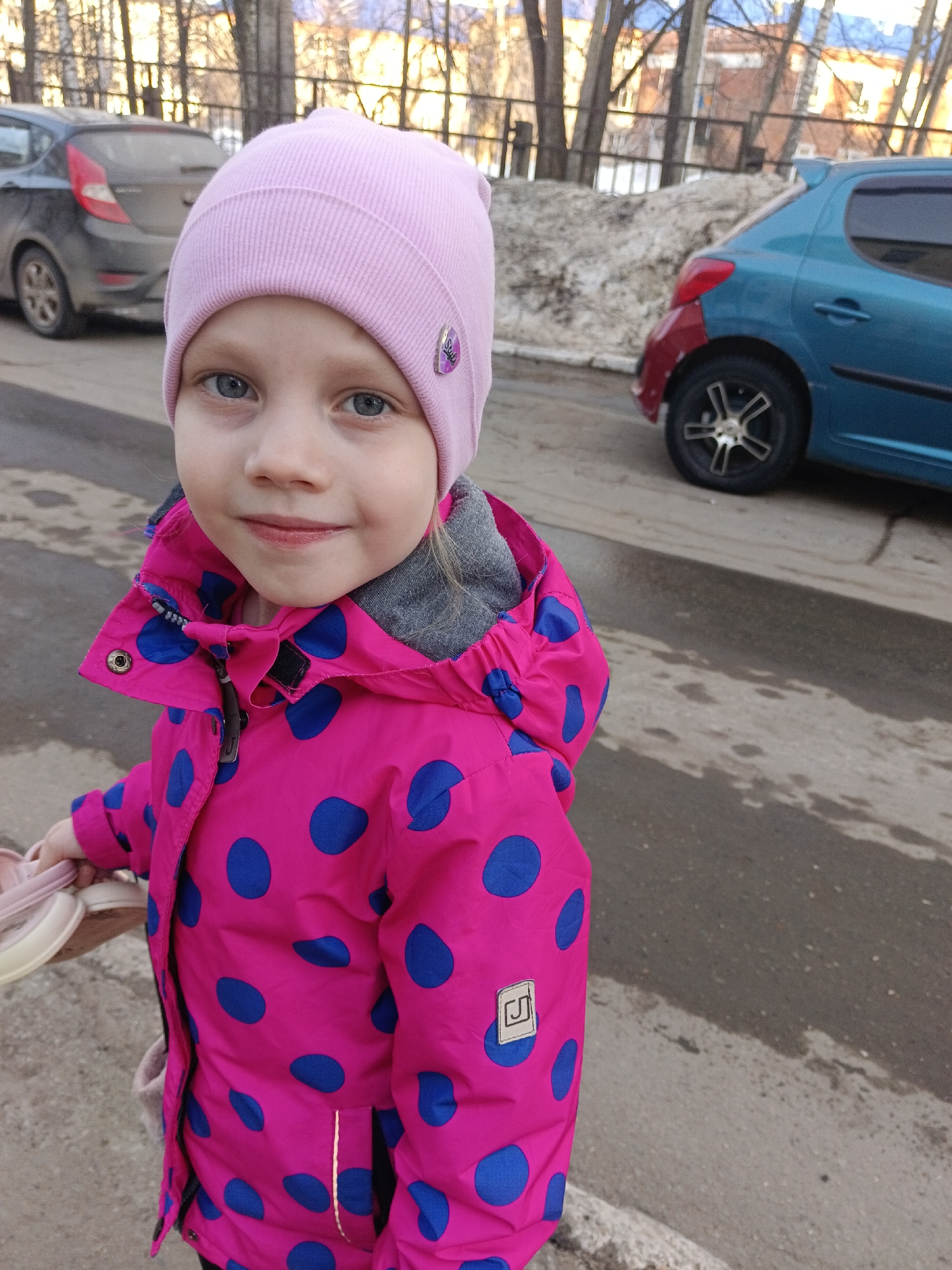 A young girl stands on an asphalt road on a partially cloudy day. She is fair-skinned with blue eyes and is wearing a pink knit hat and a pink polka dot jacket with a gray hoodie. She is holding a pair of pink shoes in her left hand. In the background, there are two cars: a dark gray hatchback and a blue hatchback with a red rear fender. Bare trees and a building are visible in the distance. Based on the appearance of the architecture and landscape, it is plausible that the location is Chaikovsky, Russia.