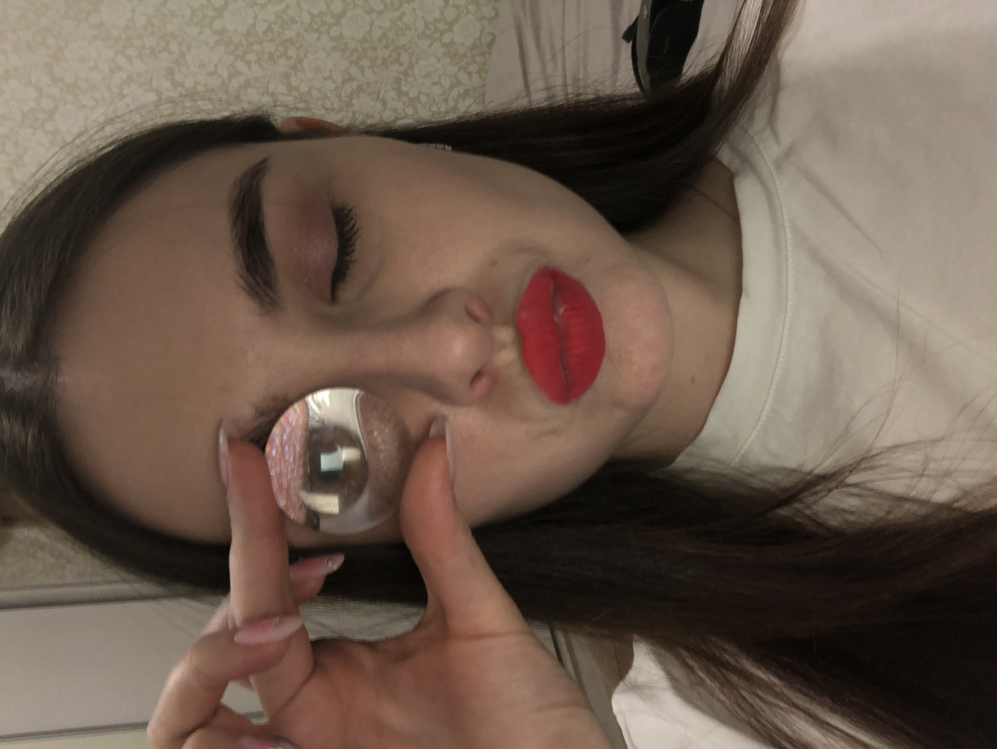 The image shows a young woman lying down, with her head tilted significantly to the left. She has long, dark brown hair that spreads around her head and rests on a white fabric surface, likely a bed. She is wearing a simple white t-shirt. Her left eye (from her perspective) is closed, and she has visible pink eyeshadow and dark mascara. Her lips are painted a vibrant, matte red and are puckered in a "kiss" shape.
With her right hand, adorned with long, light-colored, glittery manicured nails, she holds a small, clear, spherical object. This object is positioned directly in front of her right eye (from her perspective). Her magnified and slightly distorted brown eye is visible through the sphere. The object also reflects light, showing glittery pink specks within it and a bright, rectangular reflection, possibly from a window or light fixture.
The background behind her head is a light-colored wall with a subtle floral or geometric pattern. The overall scene suggests a playful and artistic self-portrait, possibly intended for social media, experimenting with visual effects. The mood appears lighthearted and expressive. No text is visible in the image. The setting is indoors, consistent with a personal space like a bedroom, in Noviy, Russia.