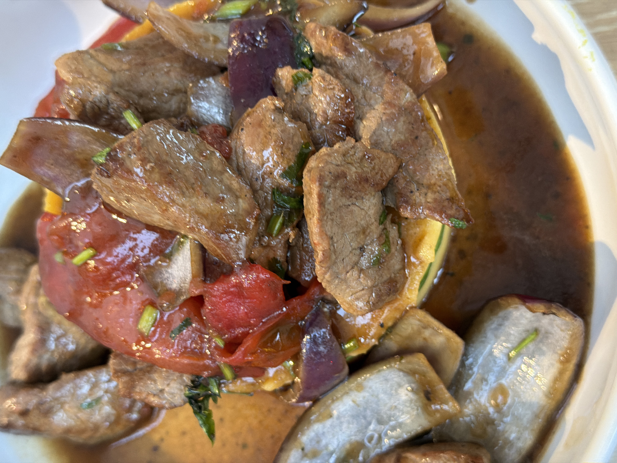 This is a close-up, overhead shot of a dish served on a white plate. The dish appears to be a stew or stir-fry featuring thinly sliced pieces of meat, likely beef, mixed with chunks of red tomato, purple onion, and possibly eggplant or bell pepper. The ingredients are coated in a glossy, dark brown sauce, with visible green herbs sprinkled throughout. The food is presented in a slightly messy, yet appetizing manner, with some pieces of meat overlapping and the sauce pooling around the components. The lighting highlights the textures and sheen of the food. There are no people or explicit environmental details visible beyond the white plate. The scene suggests a restaurant setting, possibly in Barcelona, Spain, given the context provided.
