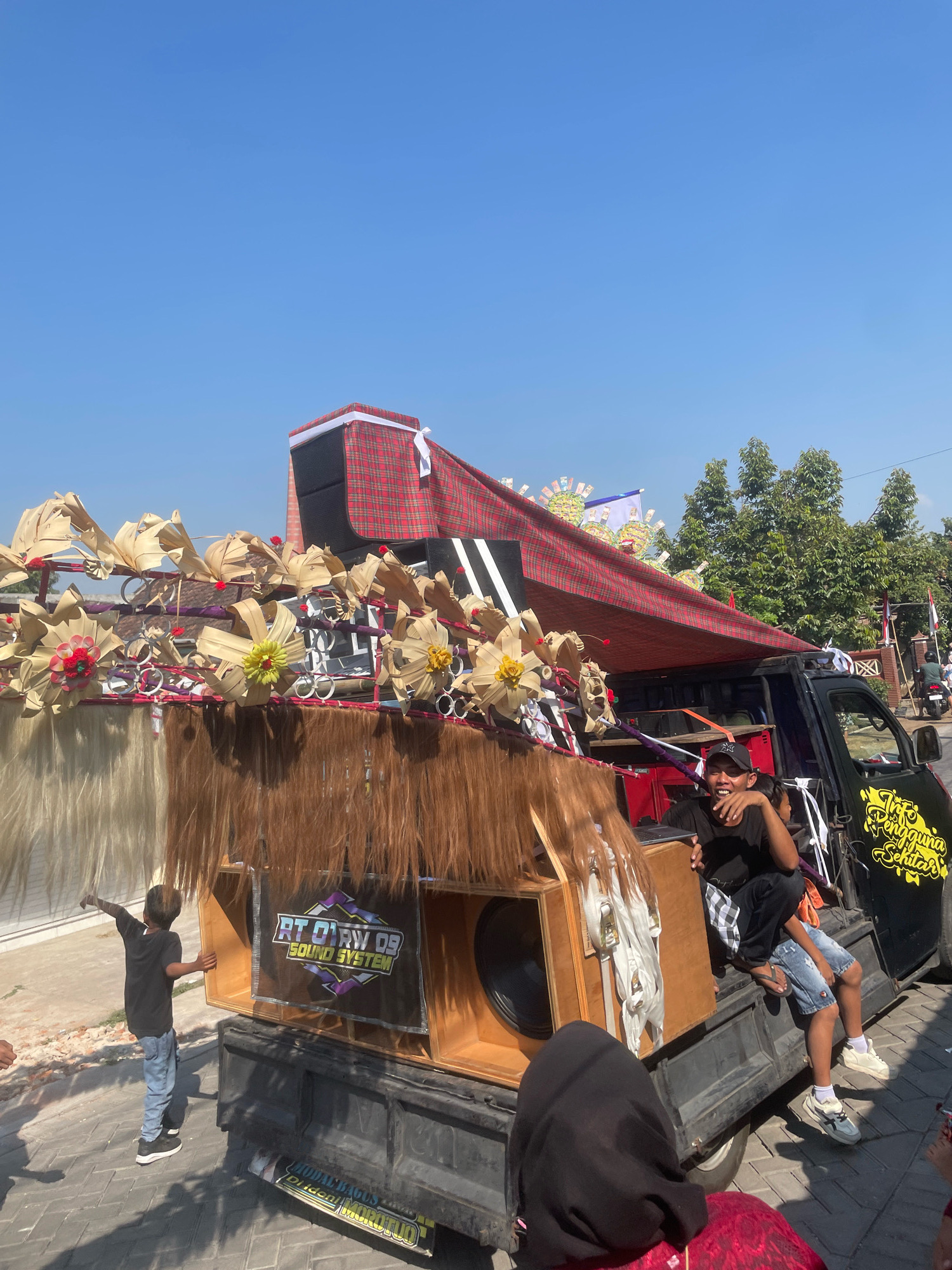 In Kebon Waris, Indonesia, a pickup truck is decorated with traditional Balinese offerings and a large sound system. The truck is adorned with woven palm leaves, flowers, and long strands of reddish-brown hair-like material. A banner on the sound system reads "RT 01 RW 09 SOUND SYSTEM". Several people are visible: a young boy in a black t-shirt and blue jeans stands on the left, pointing. Two young men are sitting in the back of the truck, one in a black t-shirt and checkered pants, the other in denim shorts. A person wearing a black headscarf and red patterned clothing is partially visible in the foreground. The truck is parked on a paved street under a clear blue sky, with trees and buildings in the background.