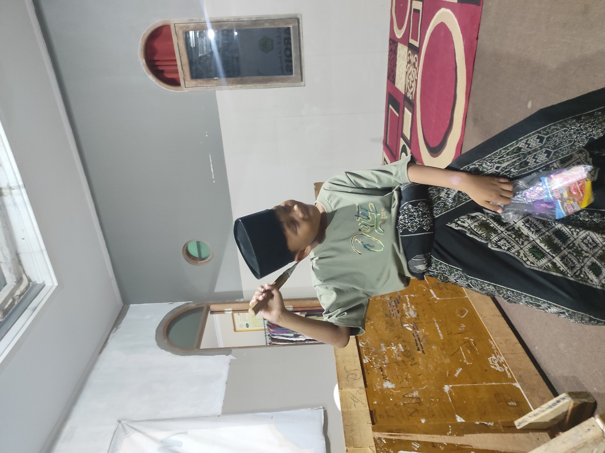 In a dimly lit indoor setting, a young boy wearing a black peci hat and a green t-shirt stands in the center of the frame. He has a piece of wood, possibly a tool, raised to his head and is looking off to the side with a slight smile. He is also holding a plastic bag in his other hand. The boy is dressed in traditional clothing, with a black and green patterned sarong wrapped around his waist. The background features a wooden partition with markings, and a patterned rug on the floor. Arch-shaped windows and a ceiling fan are visible on the walls. The overall atmosphere suggests a modest, perhaps residential or communal space, possibly in Sumedang, Indonesia, given the clothing and architectural elements commonly found in the region.