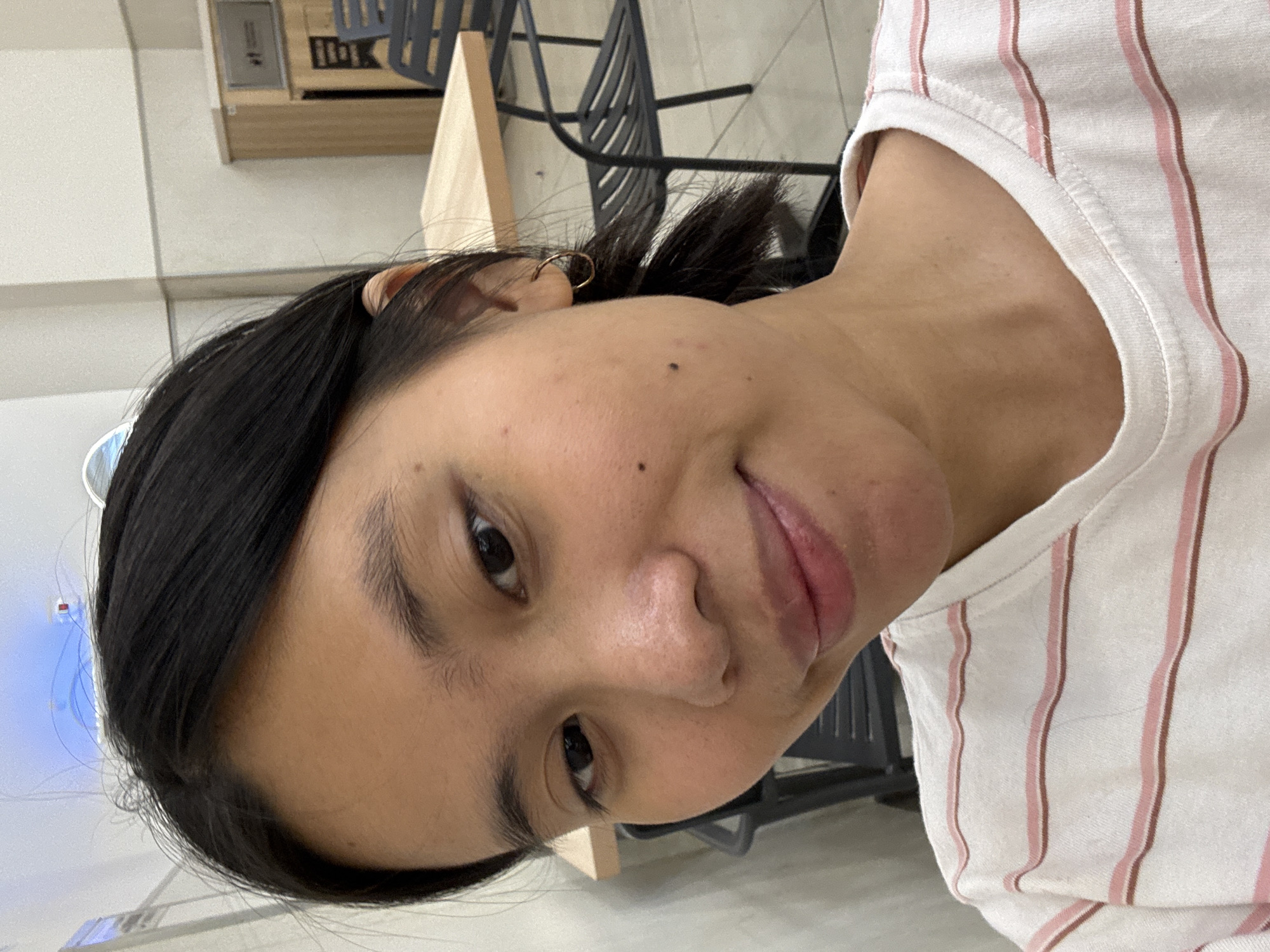 A young Asian woman is taking a selfie indoors. She has dark hair pulled back into a ponytail and is wearing a striped t-shirt. She has a small hoop earring in her left ear and a mole on her left cheek. The background shows a table and chairs, suggesting she is in a cafe or restaurant. The scene does not provide any specific visual cues to determine her precise location within East Jakarta, Indonesia.