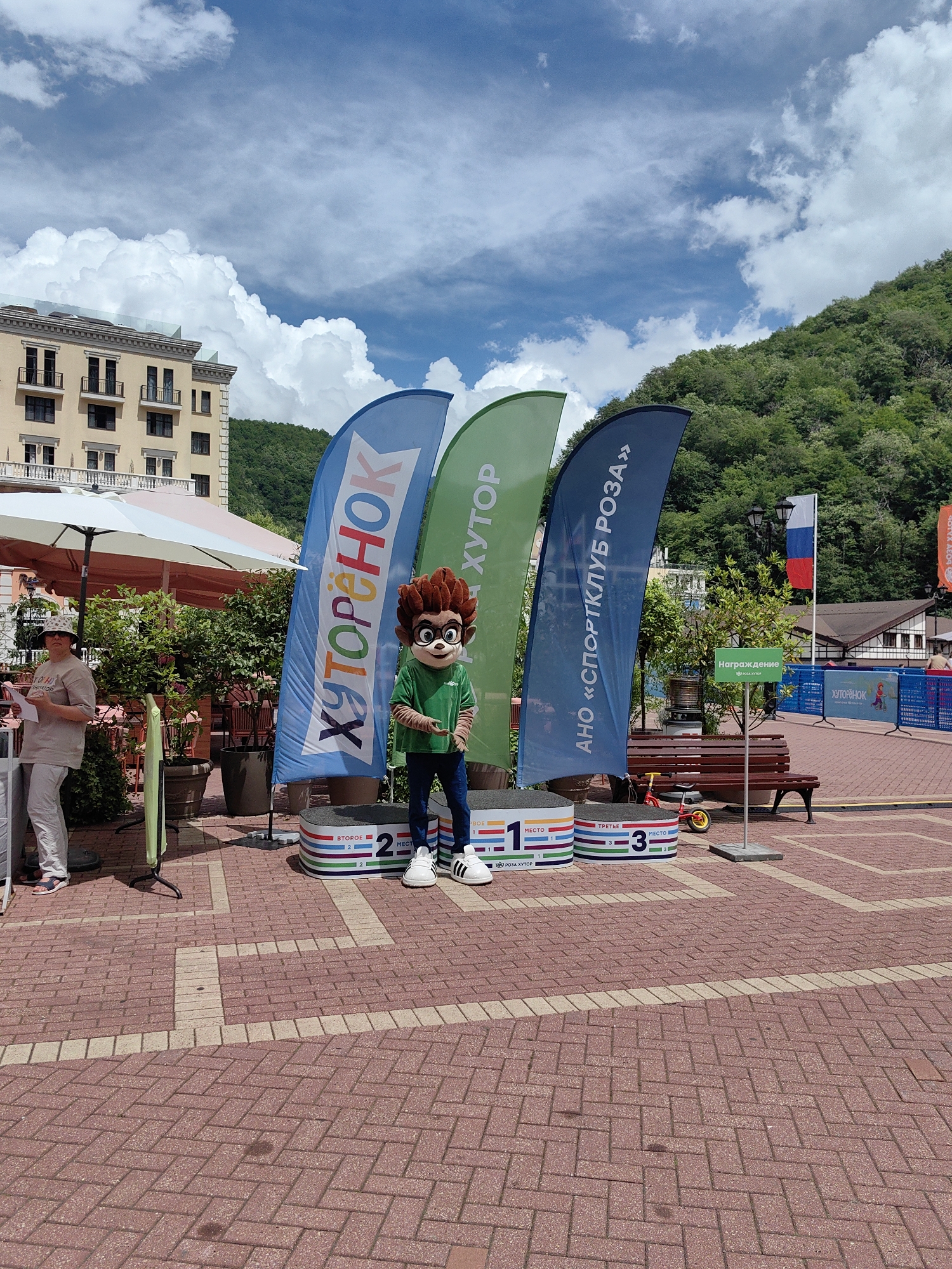 A mascot in a green shirt and blue pants stands on a podium on a brick-paved area in Estosadok, Russia. The mascot has brown, spiky hair and a round, monkey-like face with large, dark eyes. It is positioned between three large, colorful flags. The leftmost flag is blue and has white Cyrillic text that reads "ХУТОРЁНОК". The middle flag is green and also has white Cyrillic text, "ХУТОР". The rightmost flag is dark blue and features white Cyrillic text that reads "АНО «СПОРТКЛУБ РОЗА»". Below the mascot, there are three podiums labeled with numbers 1, 2, and 3, indicating first, second, and third place. A woman in a light brown t-shirt and white pants stands to the left of the podiums, holding a paper. Behind the flags and podiums, there is a building with a light-colored facade and balconies, a green hillside covered in trees, and a blue sky with fluffy white clouds. A Russian flag is visible on a flagpole in the background. A park bench and a small red tricycle are also present.