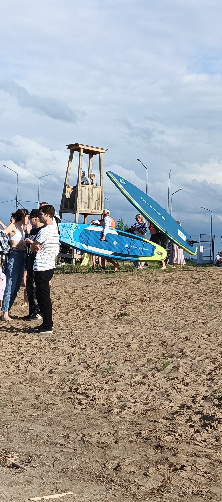 The image depicts a group of people gathered on a sandy area, likely a beach or shore, near a body of water. In the background, a wooden lifeguard tower stands tall, with a few individuals visible inside. Two large paddleboards are prominently displayed. One is blue with yellow accents and the text "AQUA MARINA" is visible. The other paddleboard, also blue with yellow accents, is being held up by a person on the right side of the frame. Several people are seen interacting, with some appearing to be waiting or observing. The weather appears partly cloudy with a bright sky, suggesting it is daytime. The overall scene suggests an outdoor recreational activity, possibly related to paddleboarding or a beach event.