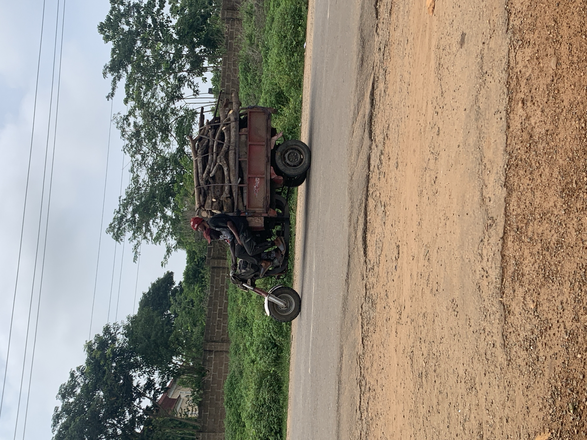The image, taken in Gbeleko, Nigeria, captures a motorized tricycle heavily laden with cut wood or firewood. The vehicle is on a road that transitions from a paved surface to a dirt and gravel shoulder on the right.
Two individuals are on the motorcycle part of the tricycle. The person operating the vehicle wears a red cap, and another person is seated behind them. The cargo bed, a reddish-brown metal structure, is packed with logs and branches of varying sizes, piled high, indicating a significant load. The motorcycle features a silver front wheel fender.
The scene is set outdoors during the daytime under an overcast or cloudy sky. Lush green trees and dense vegetation form the background, and a brick wall is visible along the roadside. Several power lines stretch across the sky in the upper portion of the image. There is no visible text identifiable on the vehicle or within the environment. The image portrays a common sight of local transportation and resource gathering in the region.