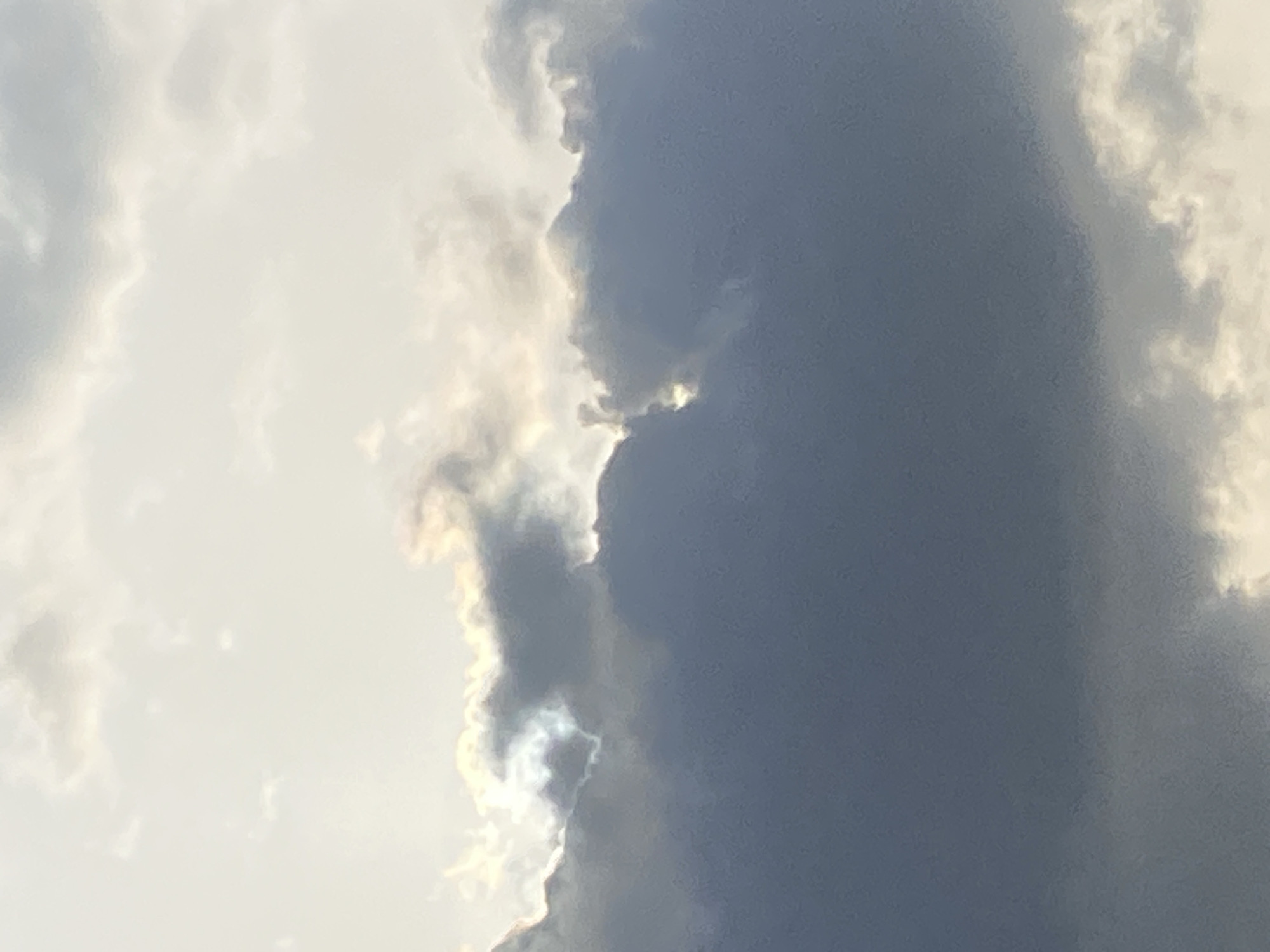 The image depicts a section of the sky with clouds. On the left side of the frame, the clouds are bright white, suggesting sunlight illumination. Adjacent to these, a large, dark gray cloud formation occupies a significant portion of the right side of the image. The edge where the bright and dark clouds meet is highlighted by refracted light. The location of the image is unknown.