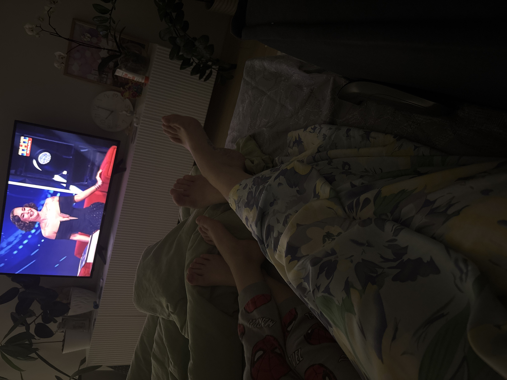 A low-angle, slightly out-of-focus indoor shot depicts a television screen on the left showing a woman in a black dress on a blue background. To the right of the television, a person's feet are visible, with another set of smaller feet appearing lower in the frame, suggesting a child is present. A patterned duvet or blanket covers the lower portion of the frame, obscuring much of the environment. The lighting is dim, and details are hard to discern. The location is possibly a residential setting. There is no concrete evidence in the image to support the claim that this image was taken in Chaikovsky, Russia. The logo "THT" on the TV screen is a clue, as it is a Russian TV channel.