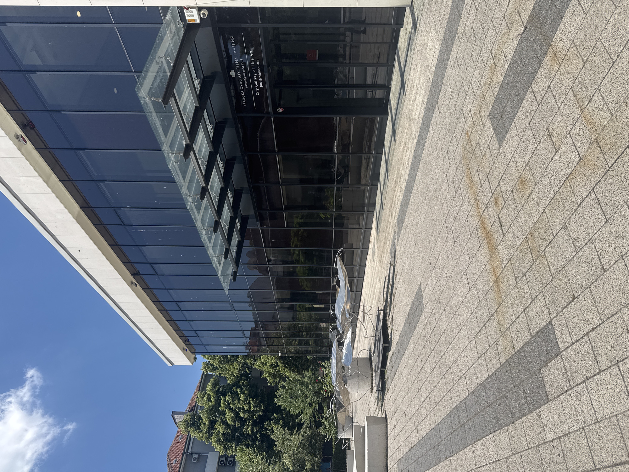 This is a daytime, eye-level, outdoor, medium shot of the City Gallery of Fine Arts in Plovdiv, Bulgaria. The building's modern architecture features a large glass facade and a white concrete overhang. A contemporary metal sculpture is situated on the tiled plaza in front of the gallery. Trees are visible in the background, as is a portion of a residential building. The sky is clear and blue with some scattered clouds.