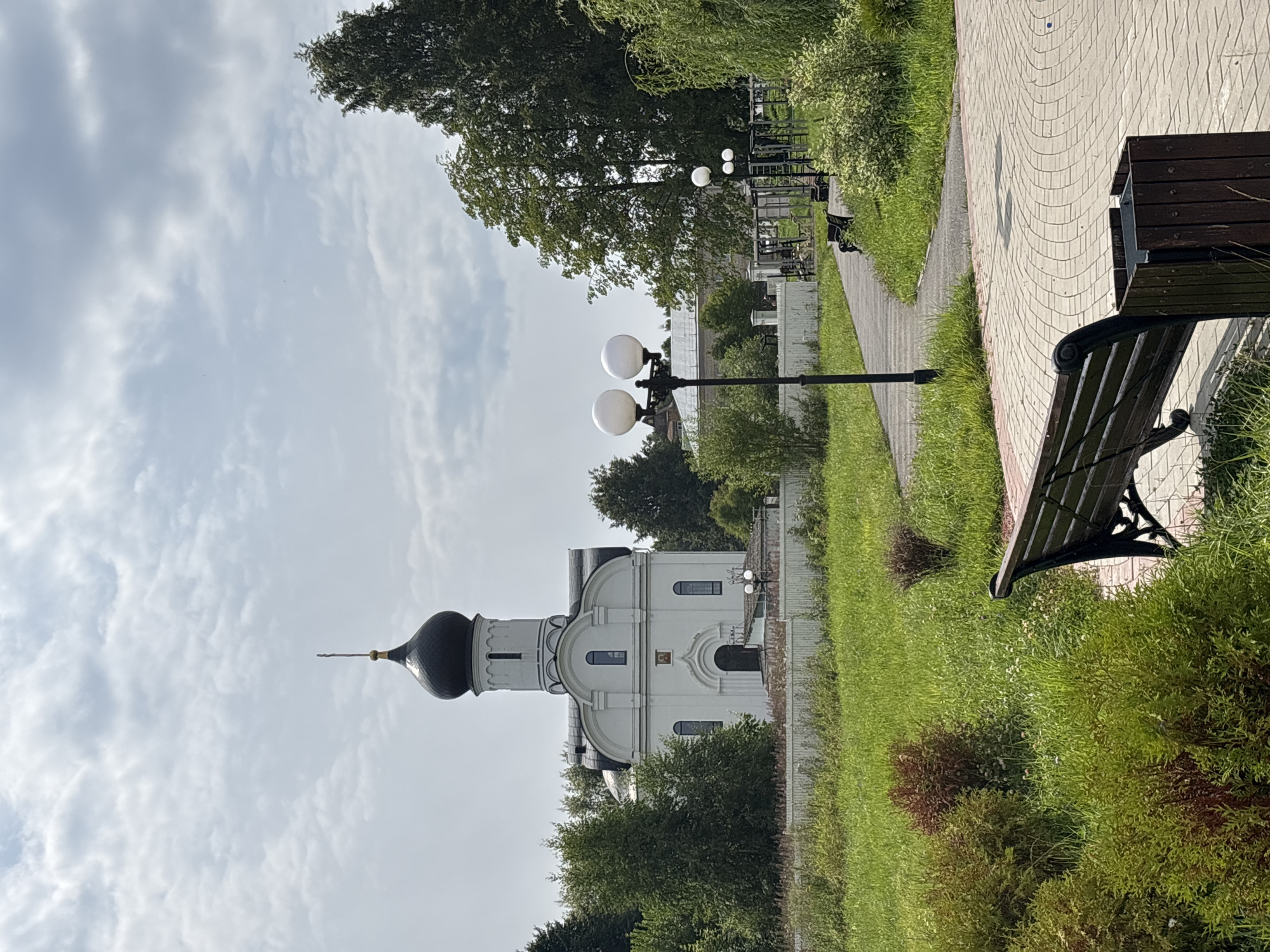 The image shows a white church with a black dome and a cross on top, set against a cloudy sky. In the foreground, there is a paved walkway with a park bench and a trash can. Lush green grass and trees surround the church and the walkway. The scene appears to be in a park or a serene outdoor area in Breytovo, Russia.