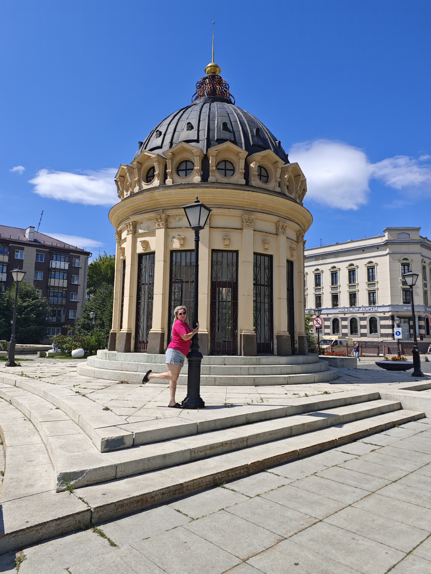 A woman in a pink t-shirt and denim shorts poses by a lamppost in front of a domed building in Kazan, Russia. The building is made of light-colored stone with a dark dome topped with a golden spire. The woman is leaning on the lamppost with one leg extended backward and her left arm resting on it. She is wearing sunglasses and black flip-flops.
In the background, there is a light-colored building with multiple windows and a sign that reads "ЭШМОКӘР ПОРТЫ" in Cyrillic. To the left of the domed building, there is a multi-story residential building with balconies. The ground is paved with concrete slabs and there are several sets of stone stairs leading up to the domed building. The sky is blue with some white clouds. The scene appears to be a public square or park on a sunny day.