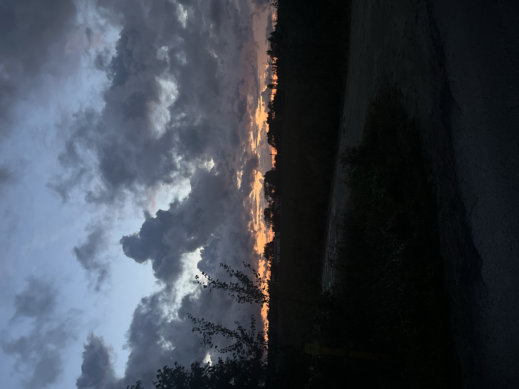 The image depicts a dramatic sunset over a rural landscape, likely in or near Vladimirovka, Russia. Dark, heavy clouds dominate the sky, with patches of vibrant orange and pink light peeking through near the horizon, indicating the setting sun. Silhouetted trees and vegetation form a dark band across the middle ground. A dirt road or path is visible in the foreground, leading into the dark landscape. The overall atmosphere is moody and atmospheric.