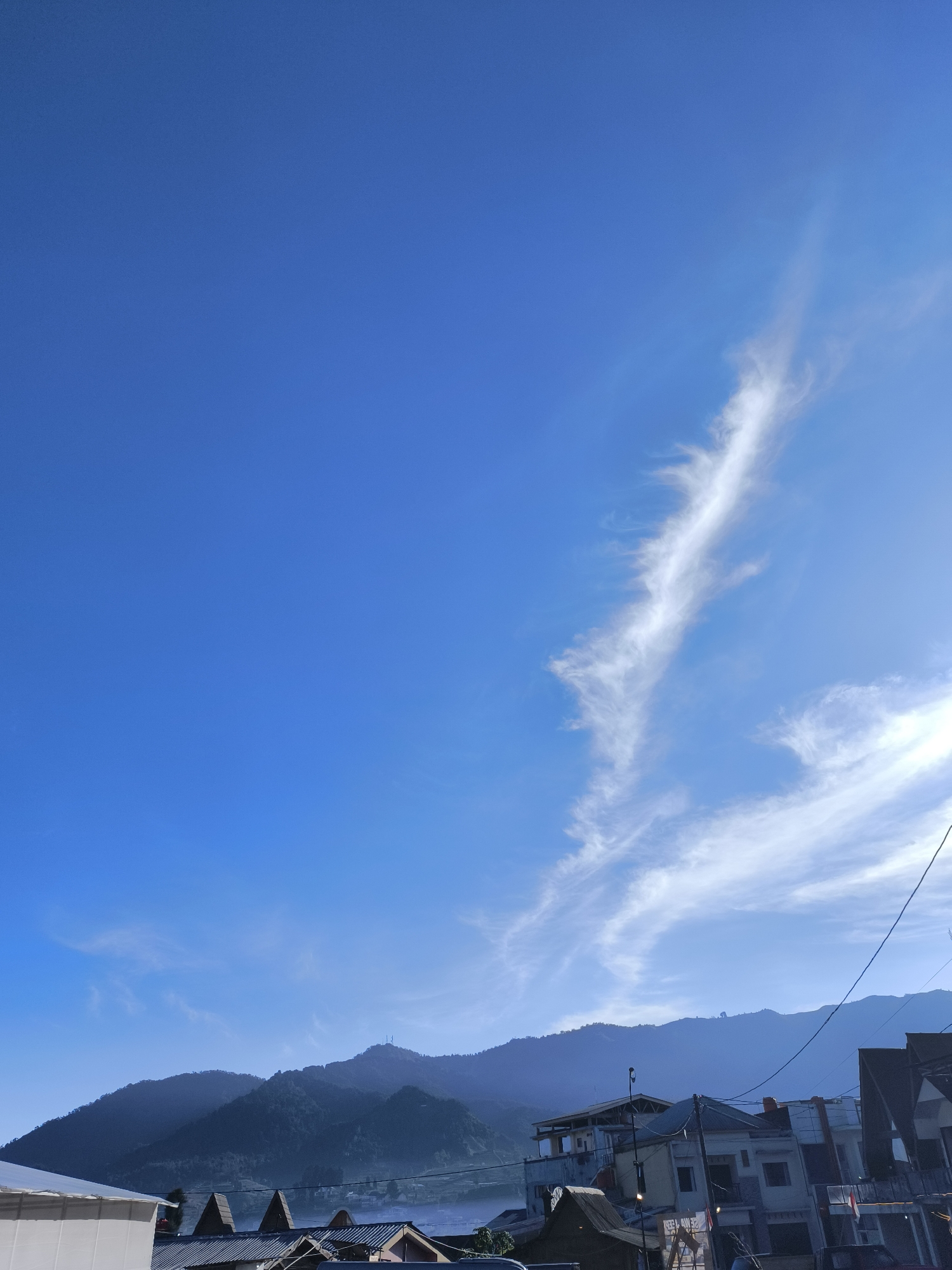 A vertical, wide-angle shot captures a clear blue sky with a wispy, elongated cloud streaking across the upper right quadrant. Below the sky, a range of dark, misty mountains forms the horizon. In the foreground, the roofs and upper stories of several buildings are visible, some with balconies and what appear to be residential structures, suggesting a populated area, possibly within Dieng Kulon, Indonesia. The light suggests it is daytime, likely morning or late afternoon due to the soft illumination of the mountains and the angle of the sun, which is not directly visible but contributes to the bright sky. There are no visible people or animals in the frame. Power lines traverse the upper portion of the buildings.