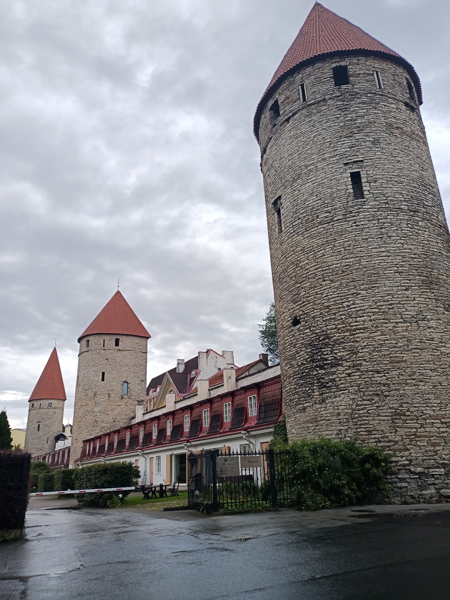 The image captures a view of the city walls of Tallinn, Estonia. In the foreground, a wet asphalt road leads to a barrier gate and a black metal fence. Beyond the fence, a building with a red roof and white walls stands alongside a stone tower with a red conical roof. To the left, another, smaller stone tower with a similar red roof is partially visible. A larger, more prominent stone tower with a red conical roof dominates the right side of the image. The sky is overcast with gray clouds. Green bushes and trees are present throughout the scene, adding to the historic and slightly damp atmosphere.