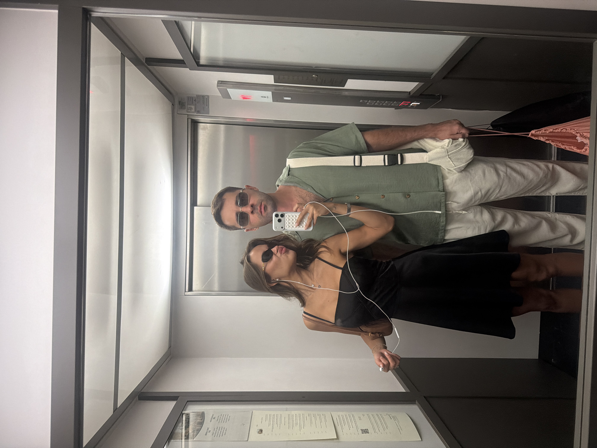Two individuals, a man and a woman, are taking a mirror selfie inside an elevator. The woman, on the left, wears a black slip dress and sunglasses, with white earbuds in her ears. She is holding a smartphone in her right hand and making a duck face. The man, on the right, is wearing a green button-down shirt, light-colored pants, and sunglasses. He has a white messenger bag slung over his shoulder and is holding a black bag with a pink drawstring bag over his arm. The elevator has metallic walls and a digital display showing the floor number. The lighting is bright, reflecting off the polished surfaces. The image appears to be taken in Verona, Italy, though there are no explicit indicators within the frame to confirm this location.