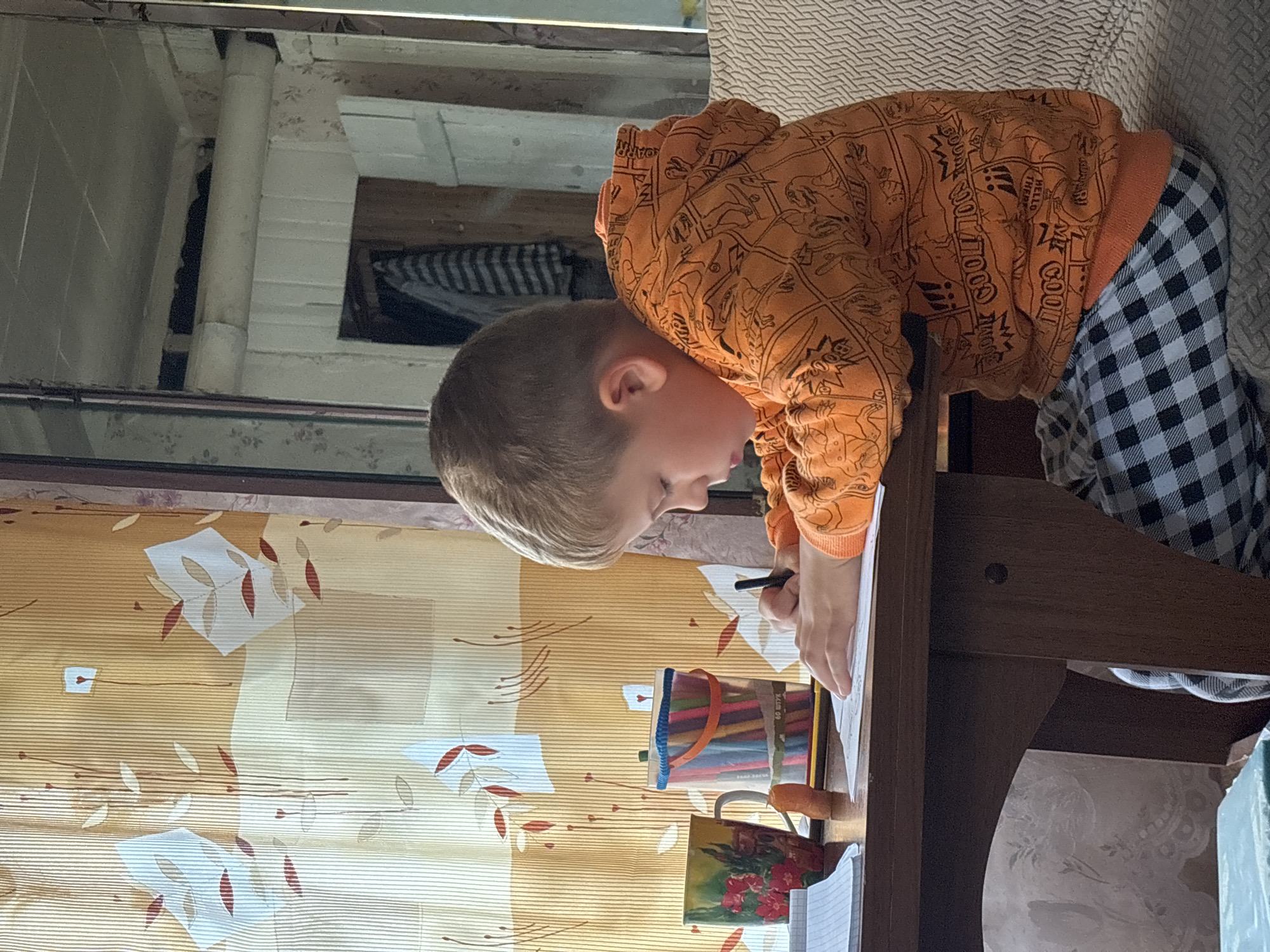 A young Caucasian boy with short blond hair is sitting at a wooden table, engaged in drawing. He is wearing an orange hooded sweatshirt with a dinosaur print and grey and white checkered pants. In front of him on the table are a notebook, a colorful mug with a floral design, a clear container filled with colored pencils, and a single carrot. The background features a curtain with a floral and leaf pattern in shades of yellow and orange, and a mirrored surface reflecting a room with wooden doors and hanging clothes. The scene suggests a domestic setting, possibly a home in Cherkasovo, Russia, given the context provided.