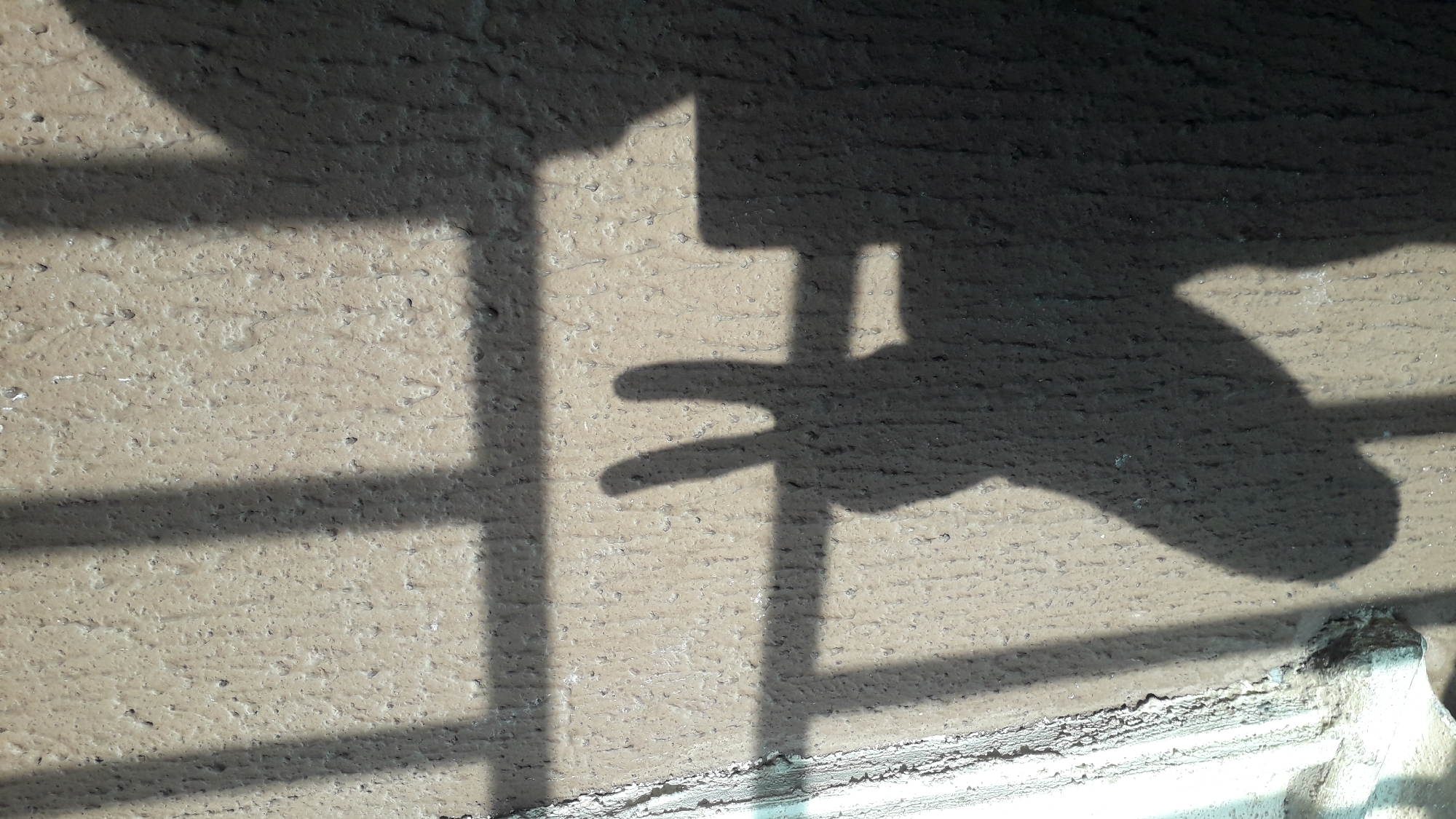 The image is a close-up, angled shot of shadows cast on a rough, beige-colored wall, likely a building exterior in Nkpor, Nigeria. A prominent shadow displays the shape of a hand making a peace sign, with the index and middle fingers extended. Behind the hand shadow, there are several vertical and horizontal shadows, suggesting the presence of a fence, bars, or a window frame. The wall surface is textured with small bumps and irregularities, possibly due to its construction material or weathering. Light appears to be coming from the left, casting the shadows to the right. There are no people or other identifiable objects visible other than the shadows and the wall itself. The overall impression is one of an outdoor urban environment.