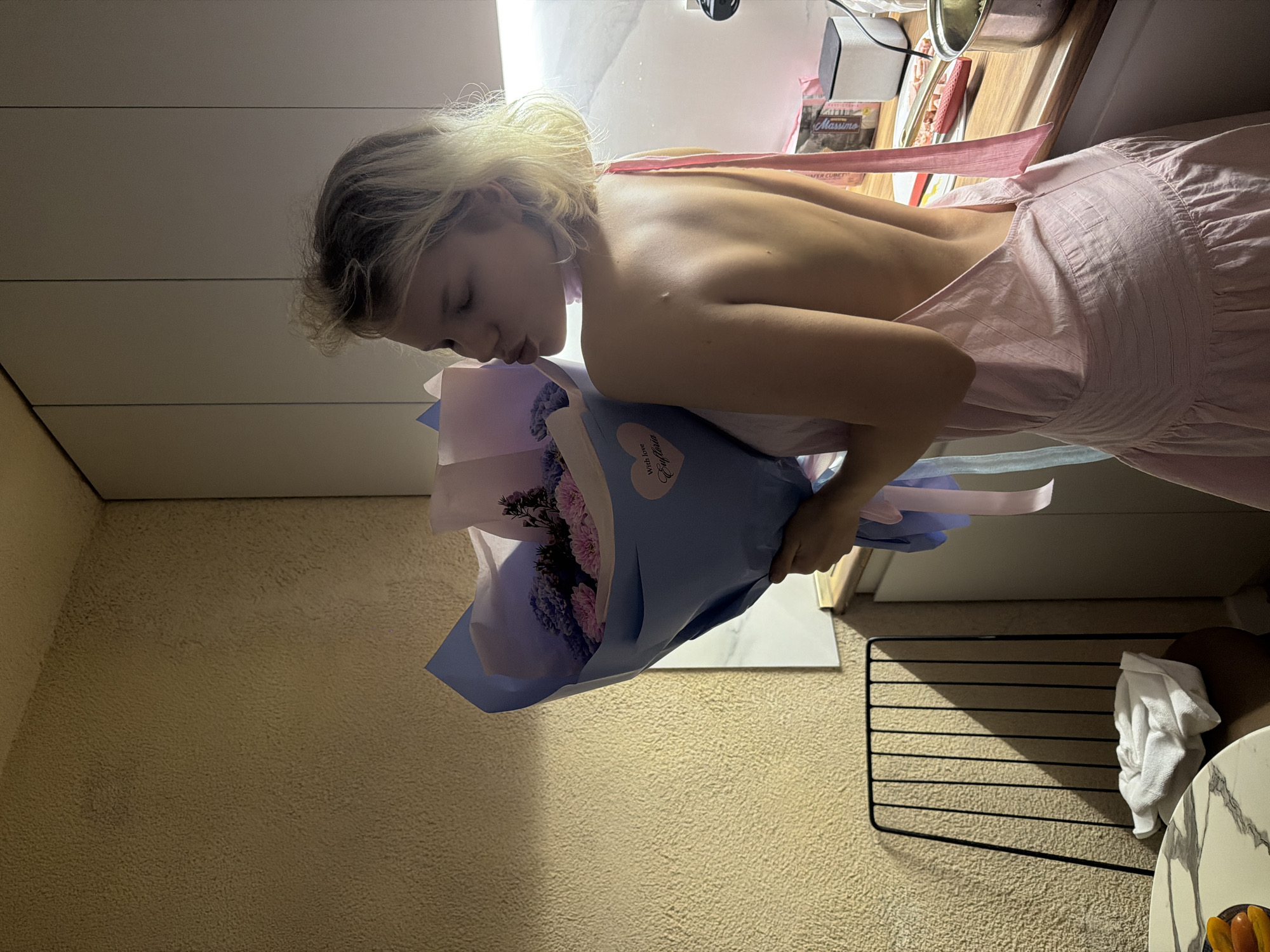 A young Caucasian woman with blonde, messy hair is shown from the back, wearing a pink, low-back dress. She is holding a bouquet of pink and purple flowers wrapped in blue paper, and she is puckering her lips. The scene appears to be a kitchen, with cabinets and a countertop visible. There is a metal chair with a white towel on it in the foreground, and a table with a marble-patterned top to the left. The lighting suggests it is nighttime or indoors. The location is not specified in the image or crops, therefore it cannot be determined if this is in Tbilisi, Georgia.